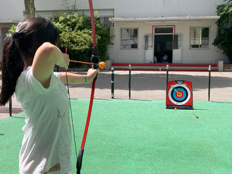 A girl with dark hair in pigtails aims a red and black bow and arrow at a target in an outdoor play area. The girl is wearing a white t-shirt, and a house with screened porch and windows is in the background. The target is on a stand, with multiple a