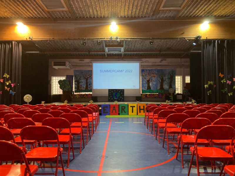 Empty auditorium with rows of red chairs facing a stage with a projection screen displaying 'SUMMER CAMP 2022'. Colorful blocks spelling 'EARTH' are positioned on the floor in front of the stage. Decorations include trees and flowers, indicating an e