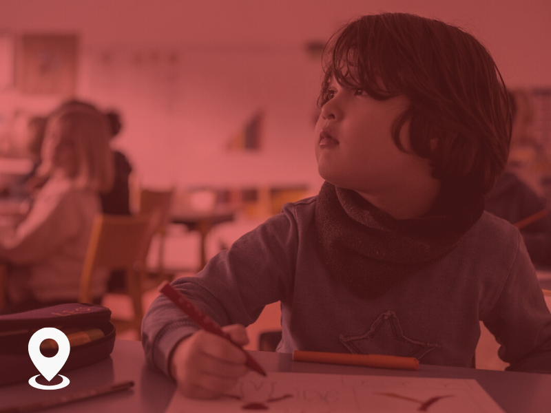 A young child sitting at a desk in a classroom, holding a pencil and working on a drawing.