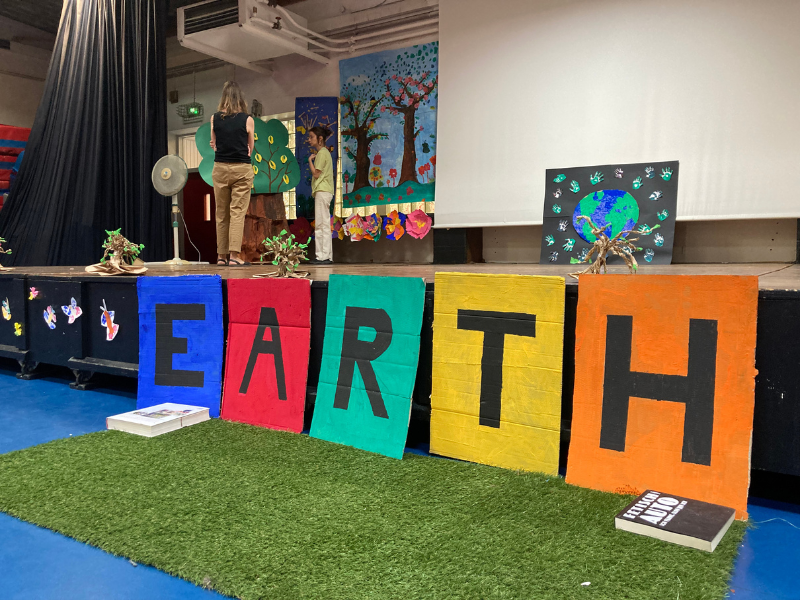 Stage with colorful signs spelling 'EARTH' in front, a large white screen, and two women preparing for a presentation. Behind them are Earth-related artwork and trees on the wall.