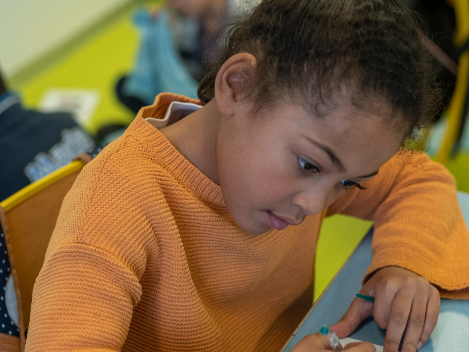 A young girl with curly hair wearing an orange sweater intently writing with a pencil in a classroom.