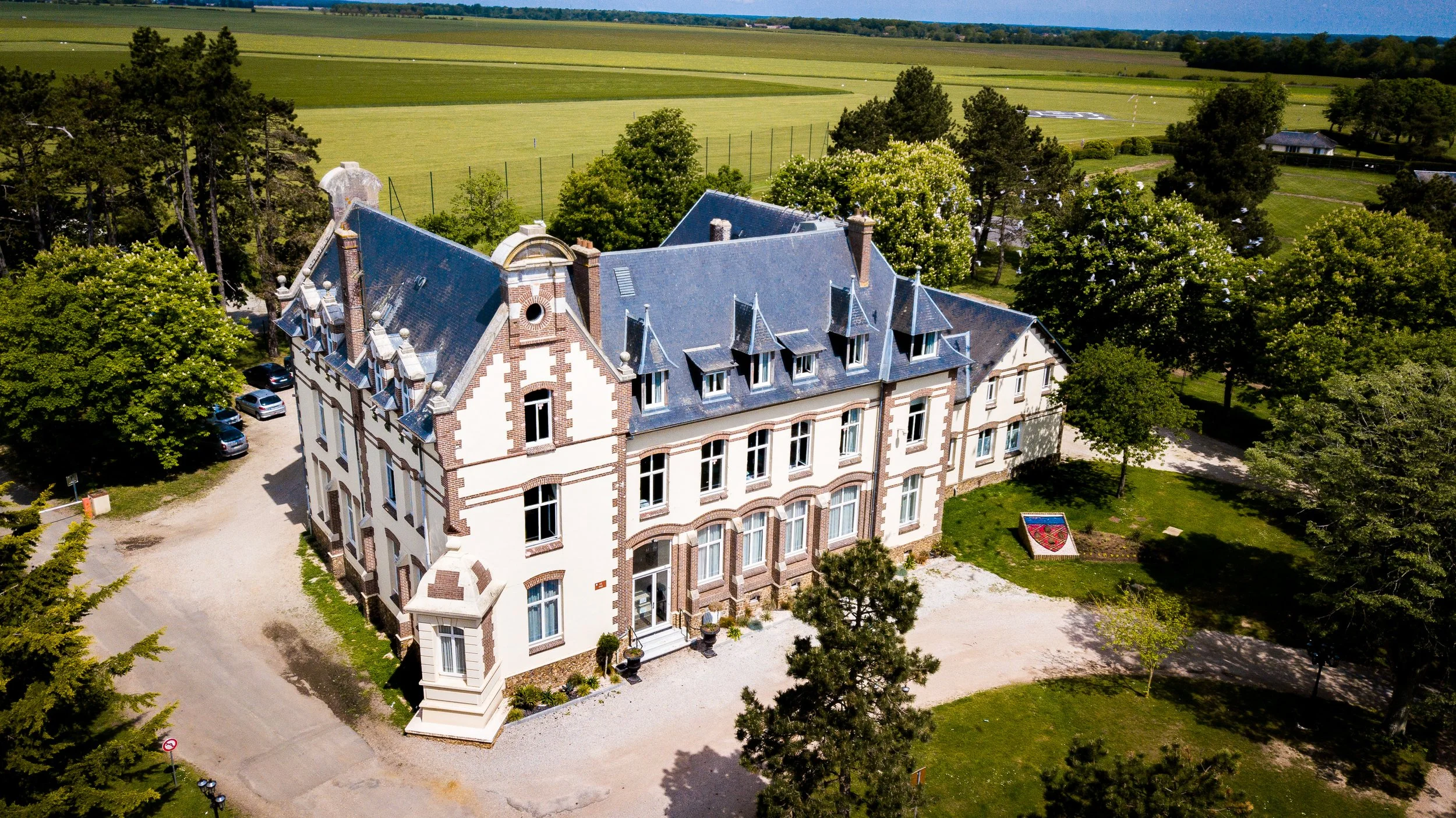An aerial view of a large, historic mansion with grey roof and white and brick walls, surrounded by lush trees and greenery, set in a rural landscape with fields and a distant horizon.