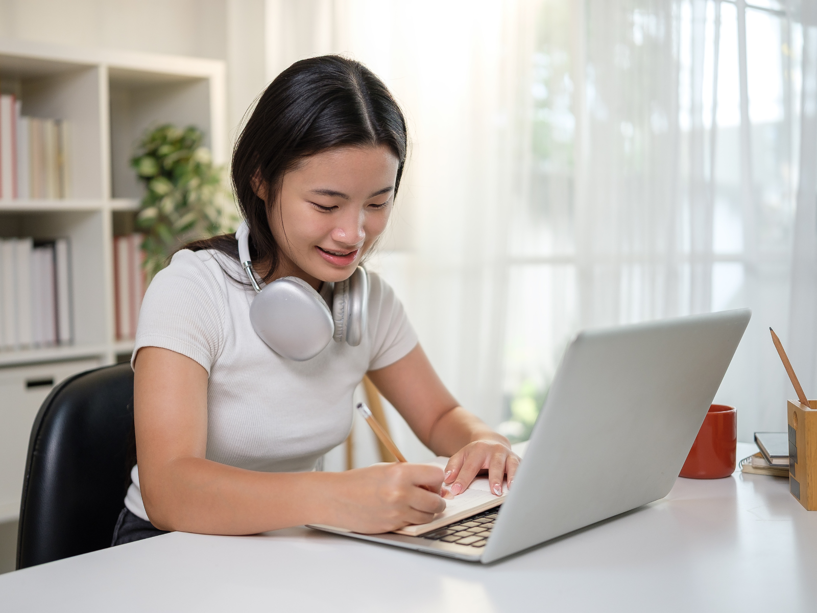A young woman with headphones around her neck is writing in a notebook while sitting at a desk with a laptop in front of her.