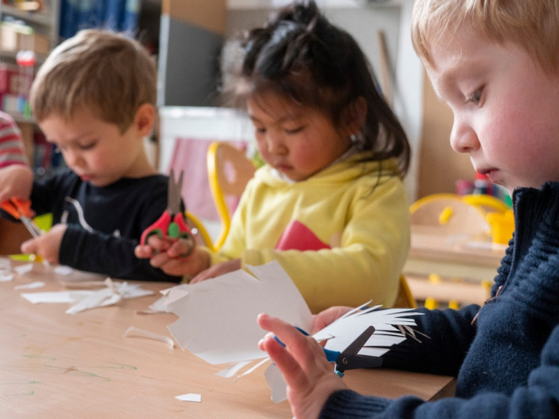 Three children sitting at a table and cutting paper with scissors, engaged in arts and crafts in a classroom.