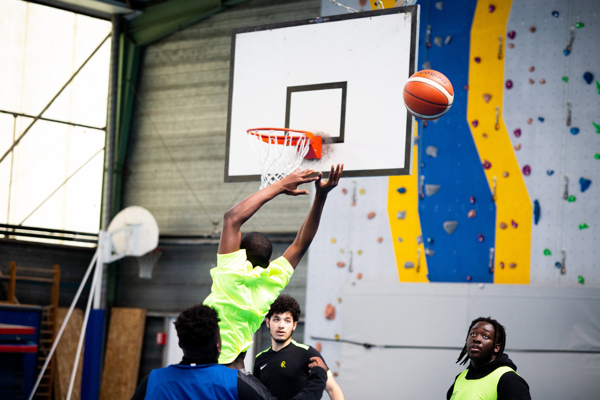 A group of young men playing basketball in an indoor gym with a climbing wall in the background. One player in a neon yellow shirt is jumping to shoot or block the ball near the hoop, with two others watching.
