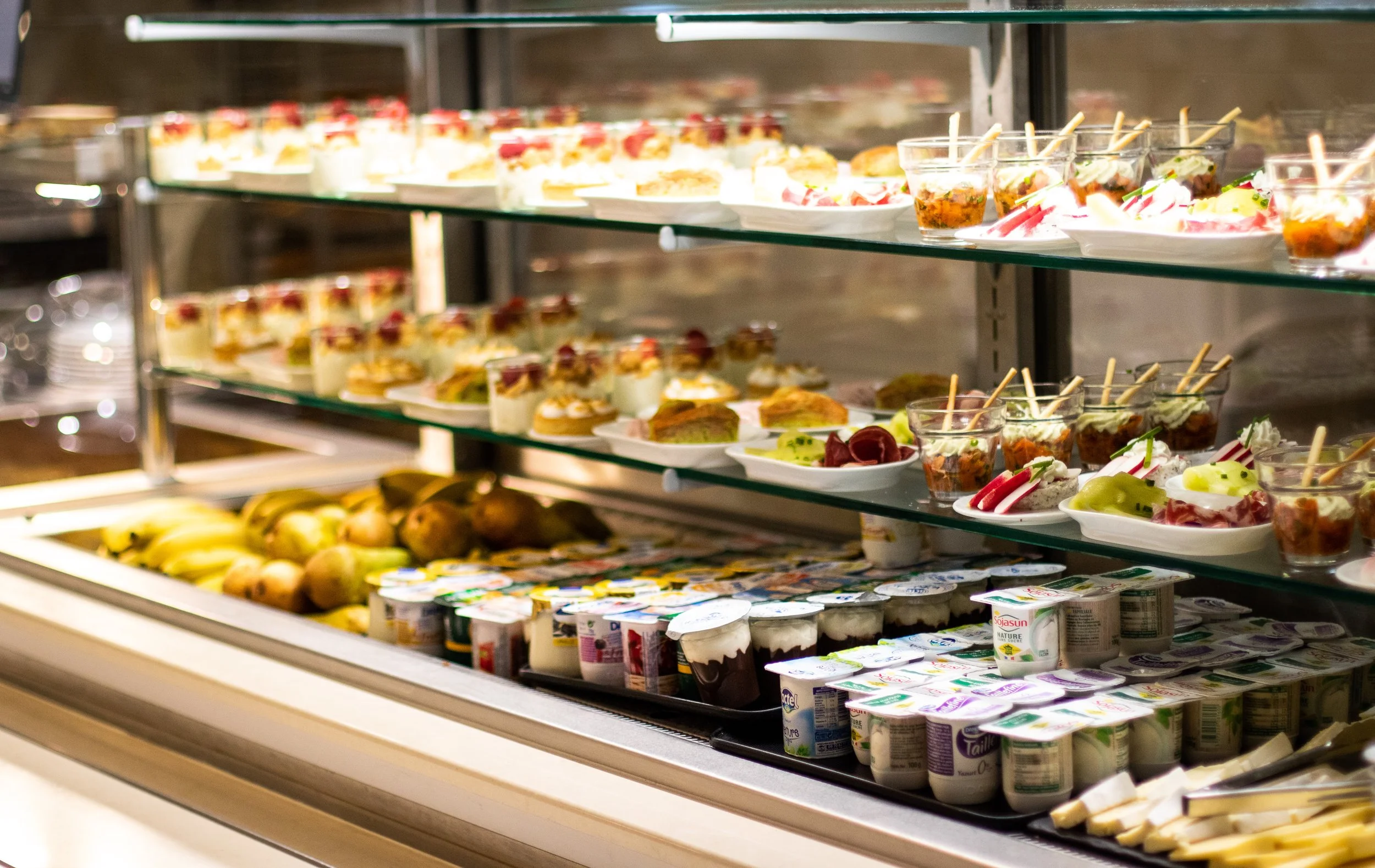 Display case with assorted desserts, fruits, and yogurt cups, including small layered cakes, cups of fruit, bananas, and packets of yogurt.