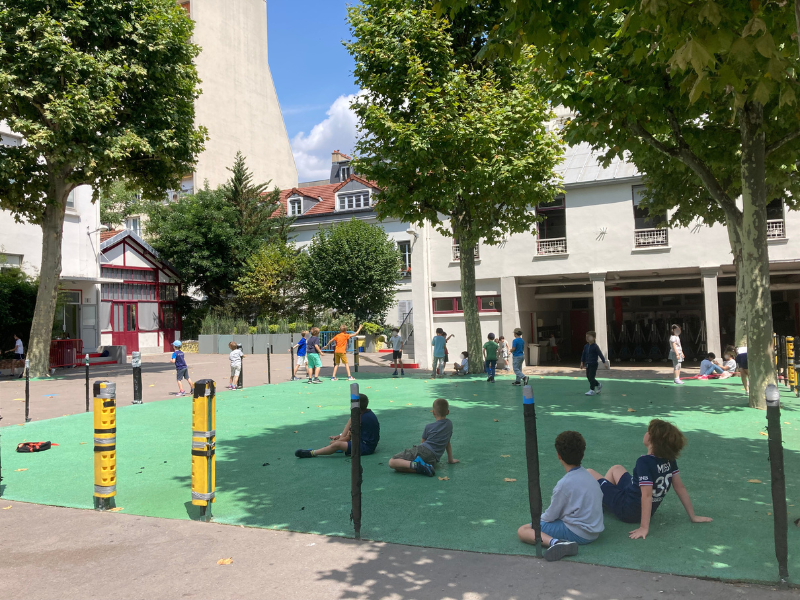 Children playing and sitting in a park with trees, surrounded by buildings on a sunny day.