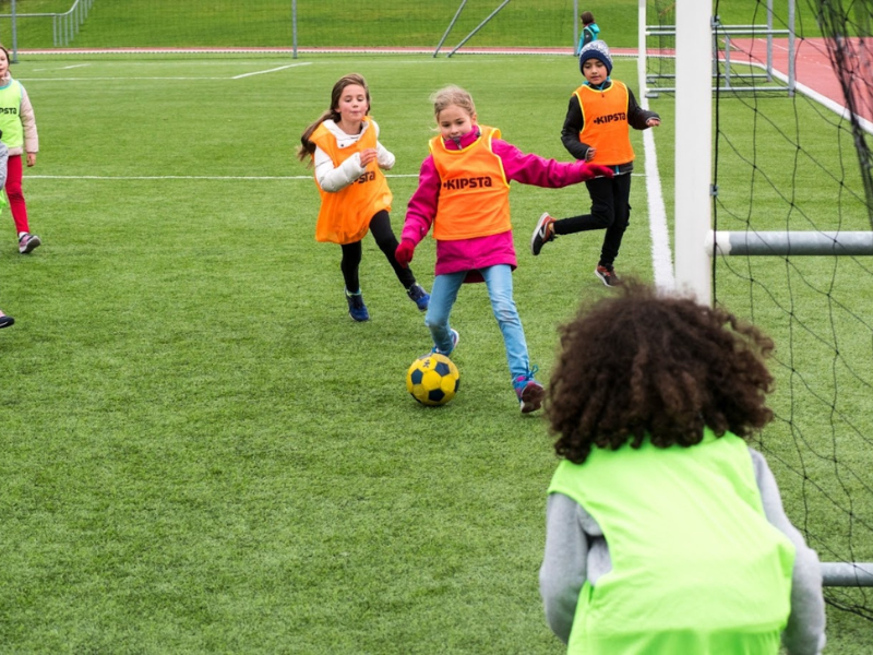 Children playing soccer on a field, with one girl kicking a yellow and black ball while others observe.