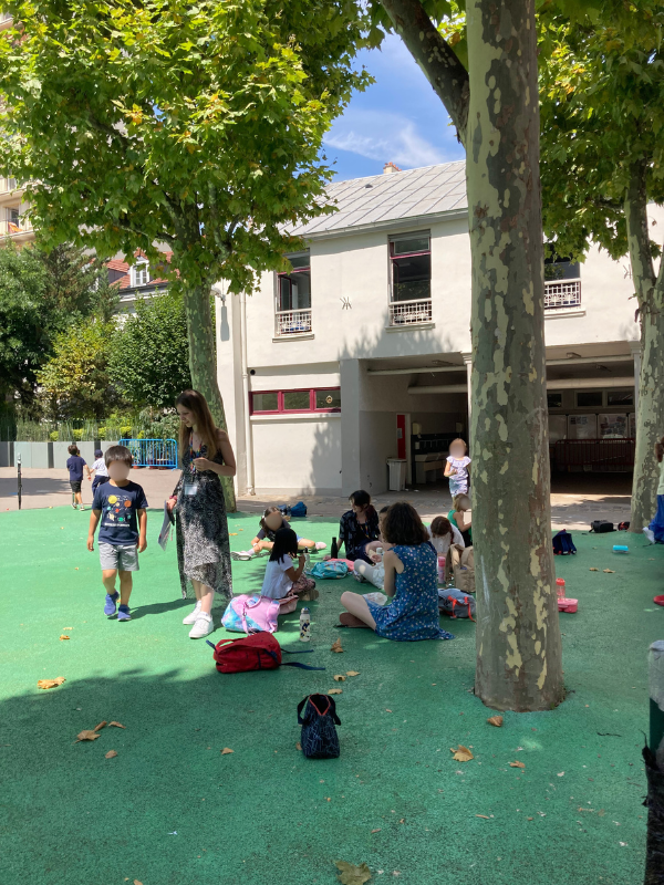 Children and adults gathering under trees in a school yard on a sunny day, with some children sitting on the ground and others walking, and a school building in the background.