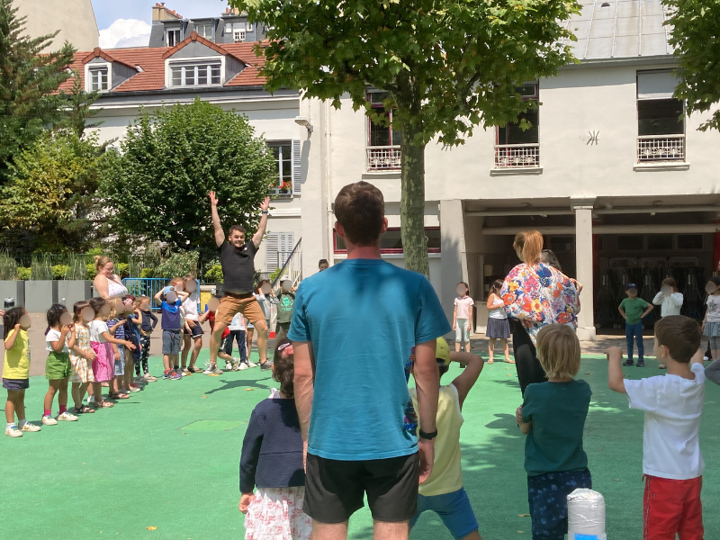 Children and adults gathered outside on a green area, with some children lined up and others watching a man jumping with arms raised, likely at a school or playground event.