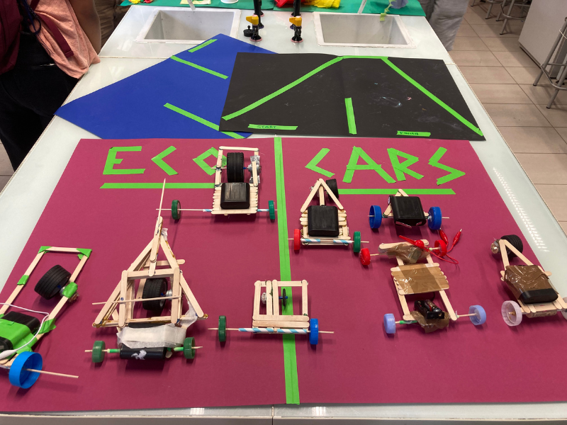 Six small remote control cars made with wooden sticks and plastic parts on a table with a sports court design and a sign that reads "ECO CARS."
