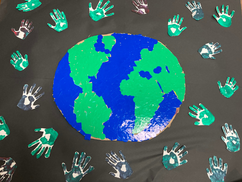 A paper collage of the Earth with green and blue sections on a black background, surrounded by cut-out handprints in various shades of green, teal, purple, and white.
