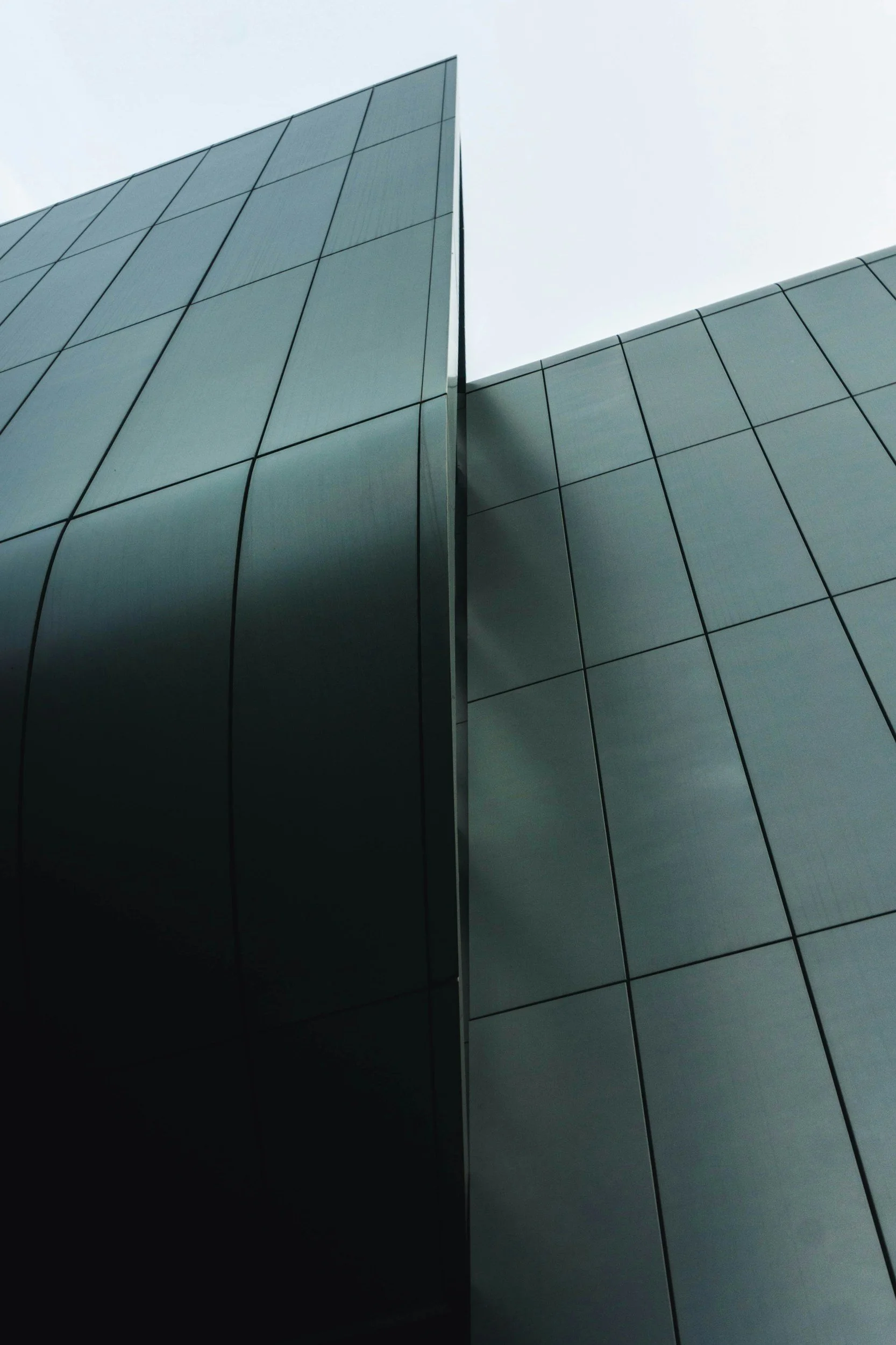 Close-up of modern building with sleek, dark gray panels and sharp angular lines, viewed from below against an overcast sky.