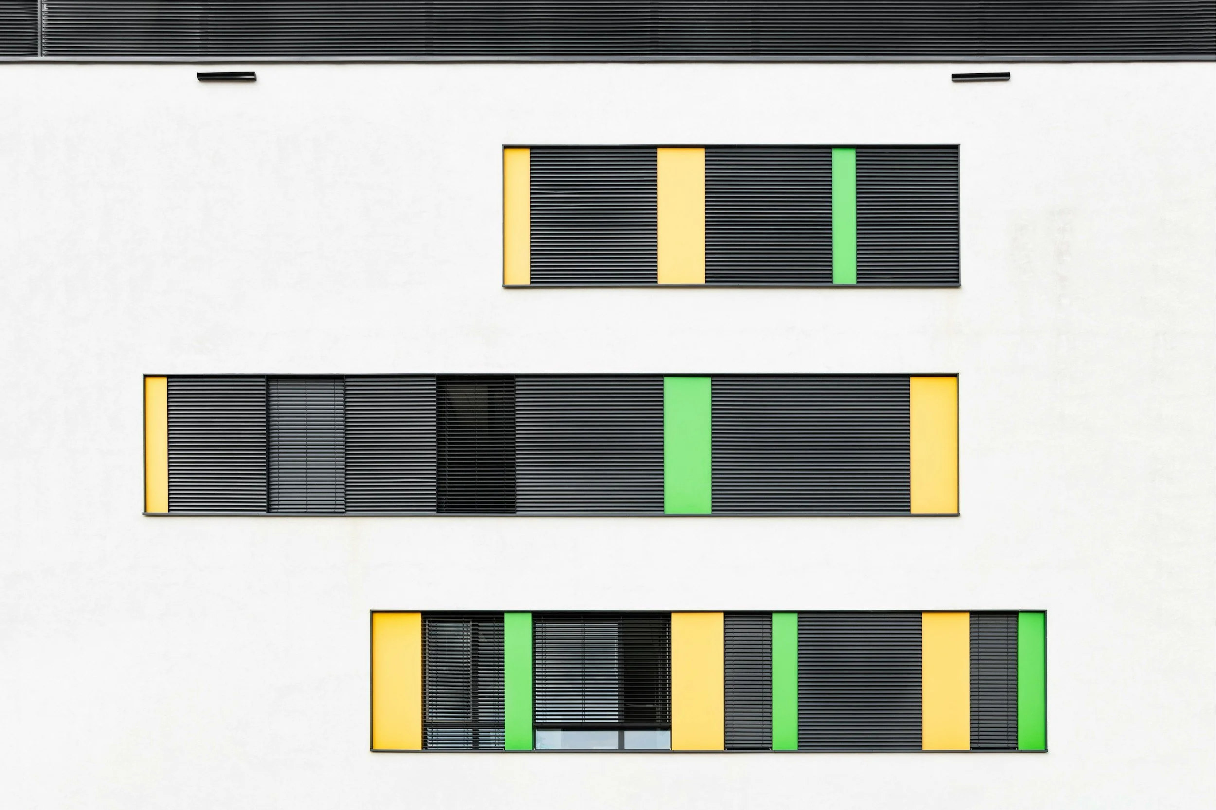 Modern building facade with three floors, featuring rows of black horizontal slats and colored panels in green and yellow.
