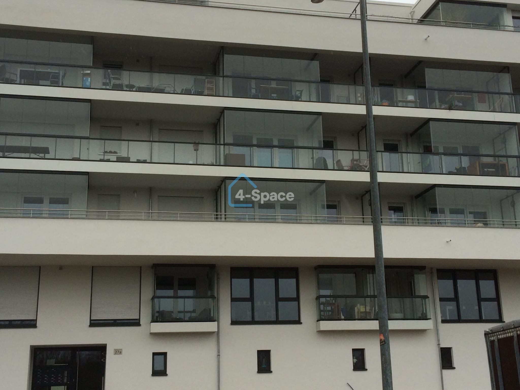 A multi-story modern apartment building with glass balconies and large windows. There is a streetlight pole in front of the building, and the photo is taken during the daytime.