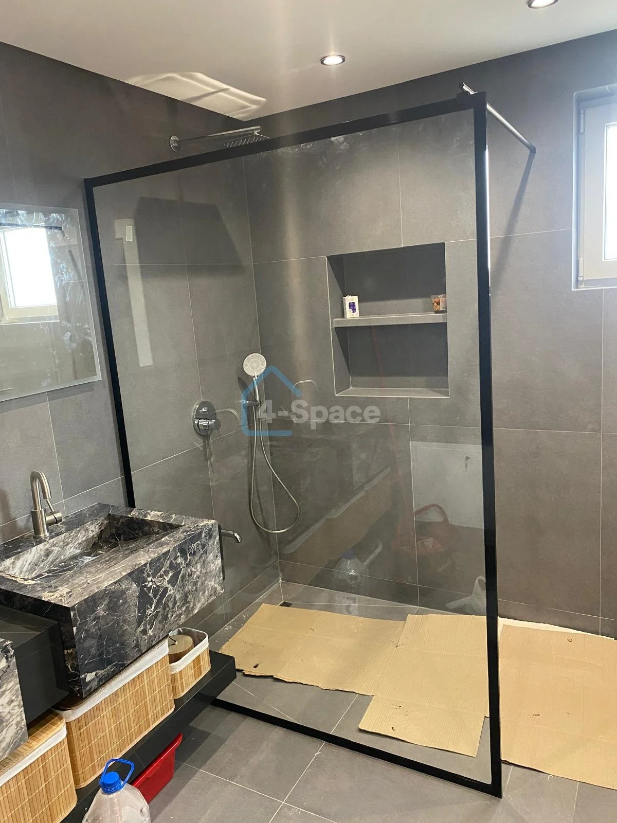 Modern bathroom with a glass shower enclosure, gray tile walls, a black marble sink, and small windows letting in natural light.