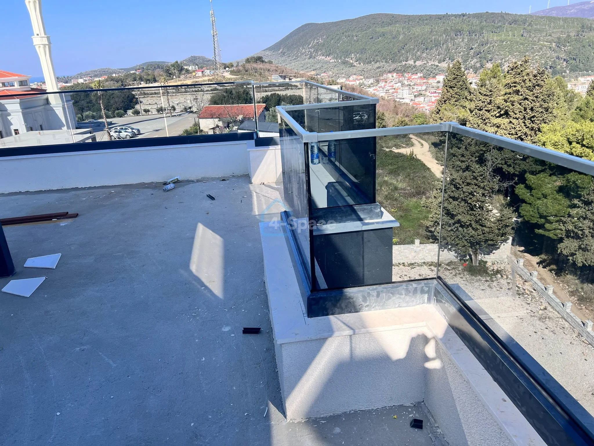 View from a rooftop balcony with glass railings overlooking a hilly landscape with trees, a dirt path, and a residential area in the distance.