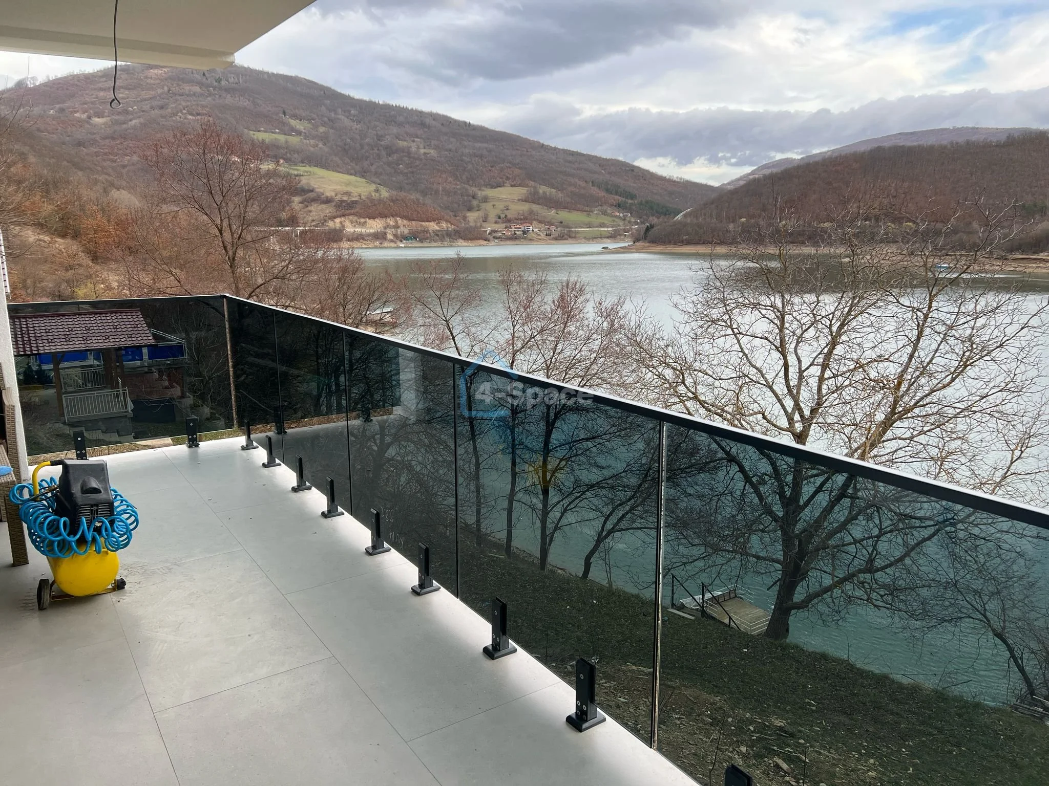 Balcony with glass railing overlooking a river and rolling hills with trees, some bare, and cloudy sky.