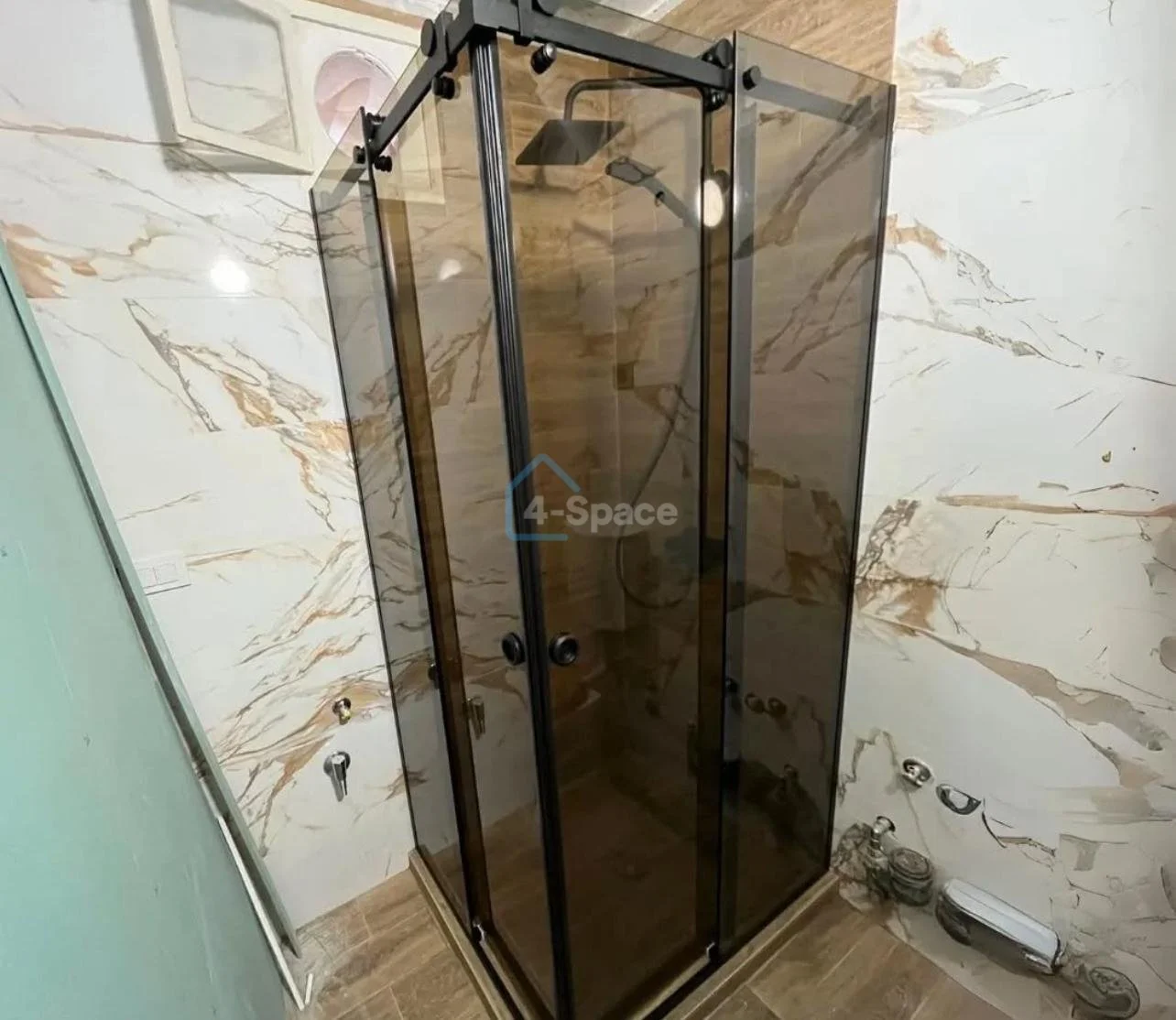 A corner shower enclosure with black framing and tinted glass panels in a bathroom with marble-patterned walls and wood-look floor.