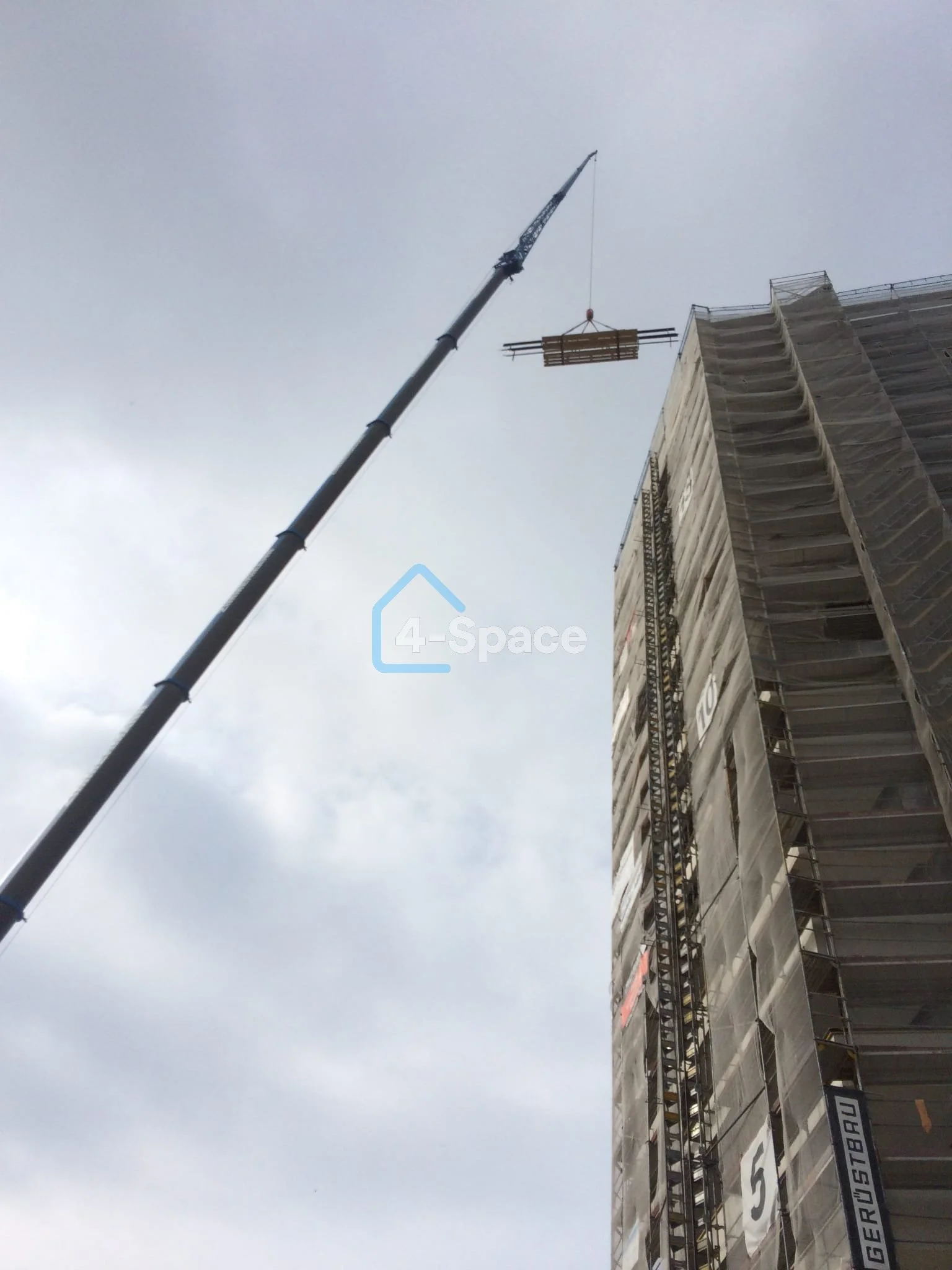 A tall construction crane lifting a load of construction materials towards a building under construction, with scaffolding on the building and a cloudy sky in the background.