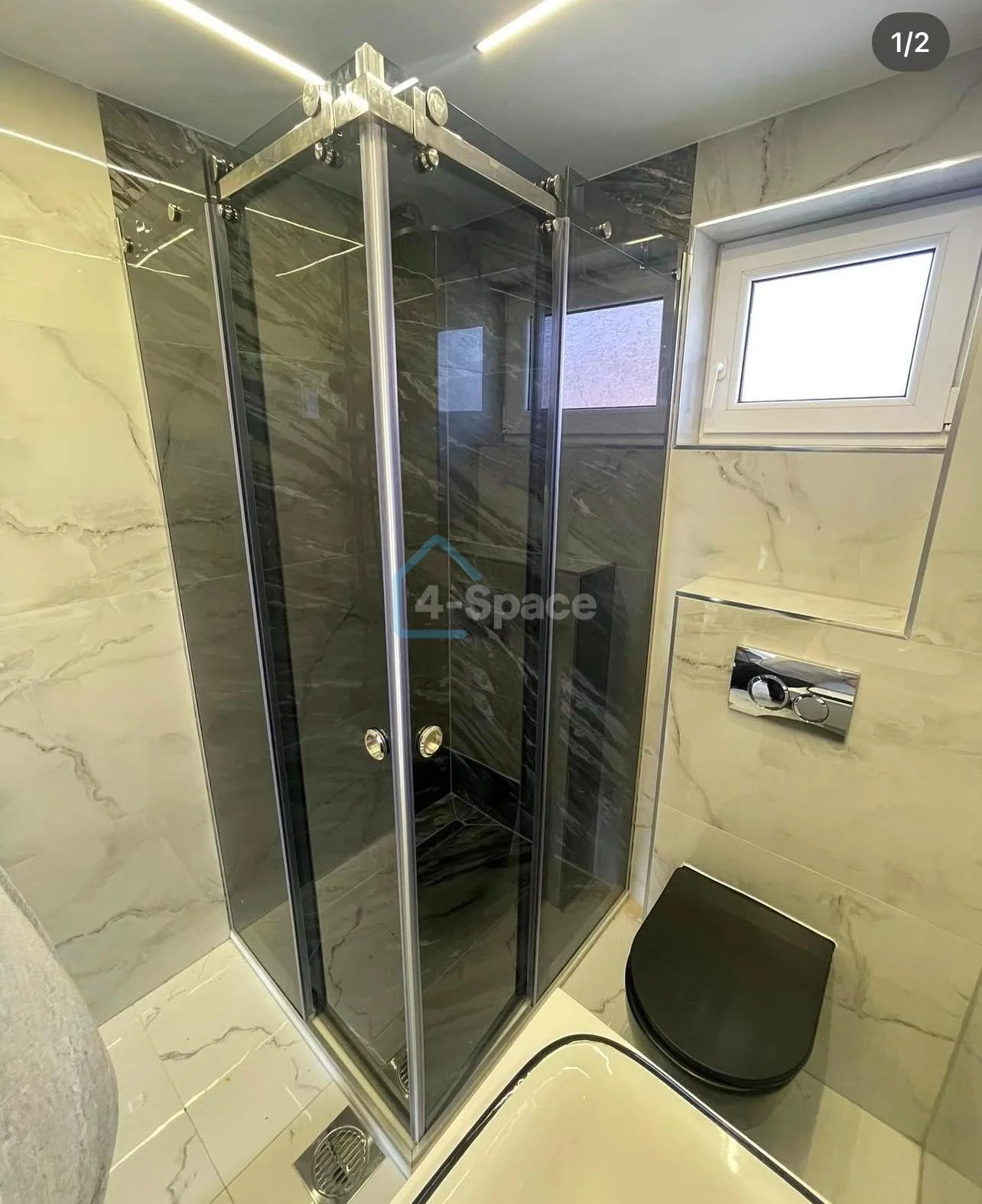 Small modern bathroom with a glass shower enclosure, black toilet, beige marble walls, and a small window.