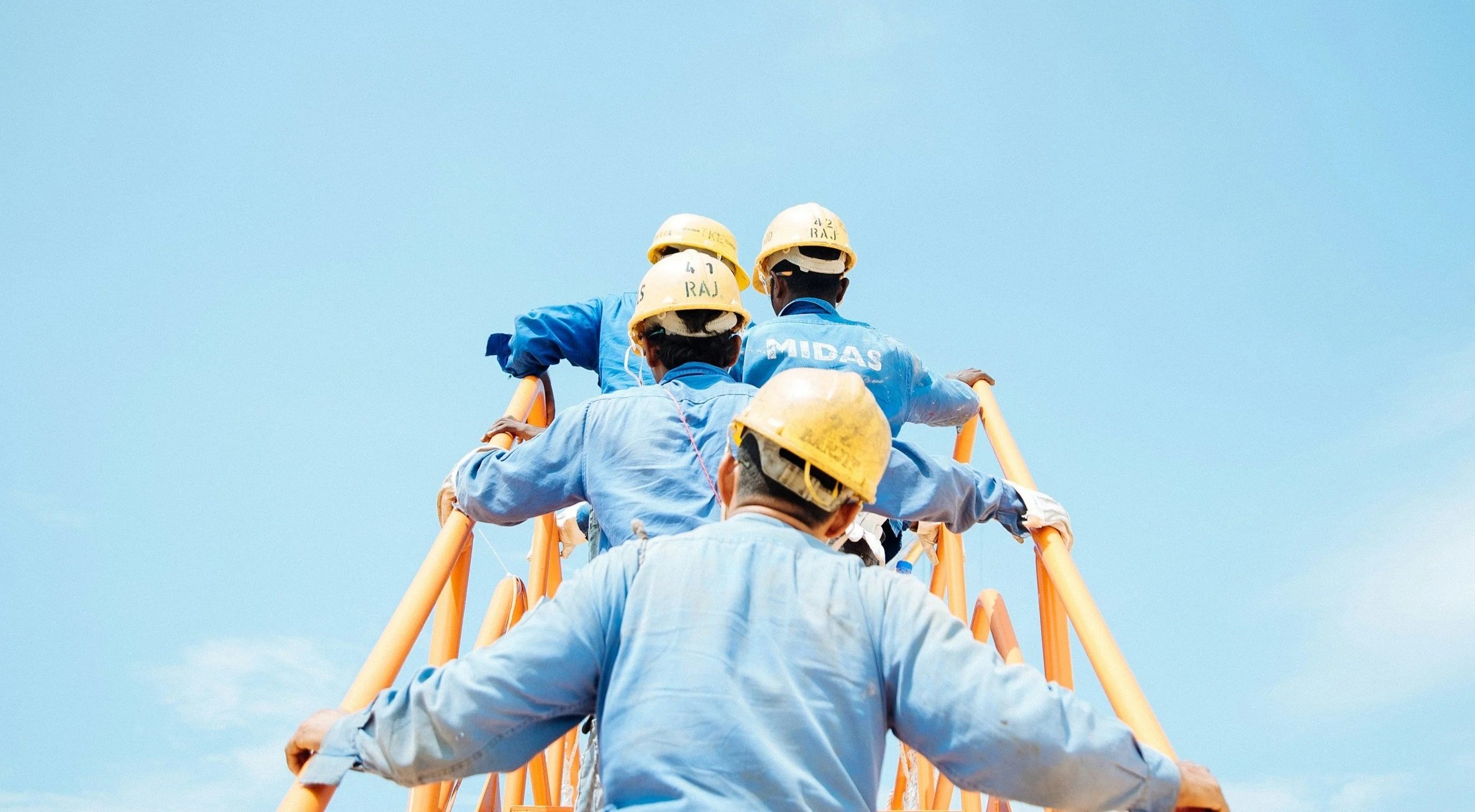Five construction workers wearing yellow safety helmets and blue uniforms climbing a tall orange ladder against a bright blue sky.