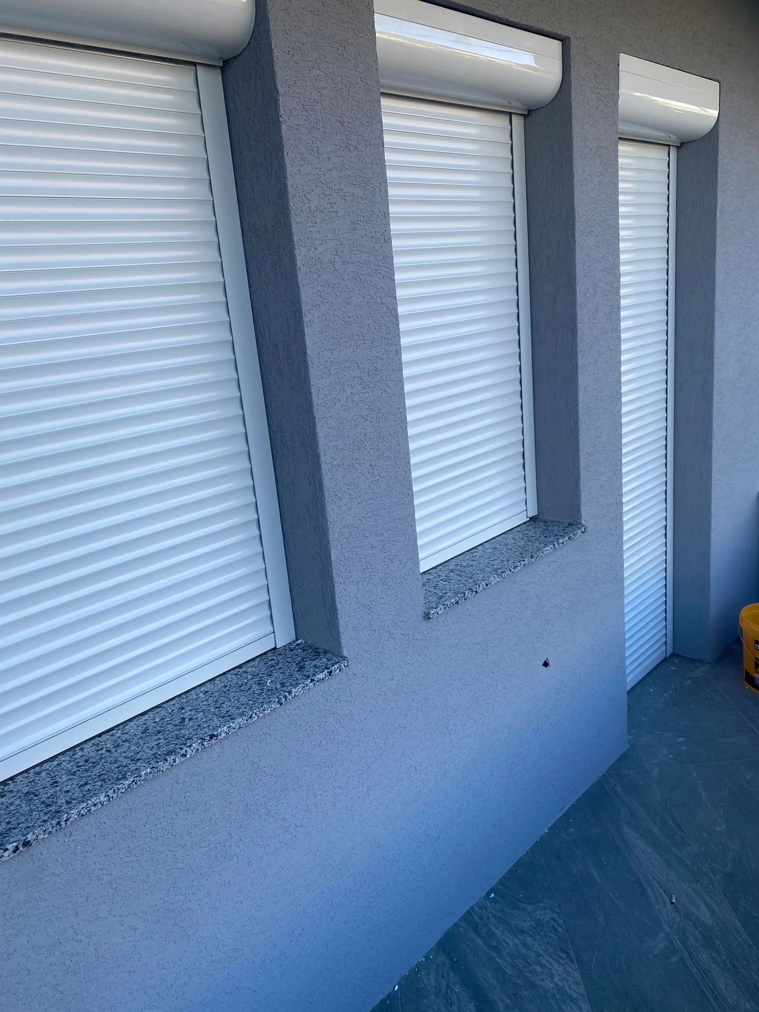 Closed white roller shutters on a gray building exterior with granite window sills and tile flooring.