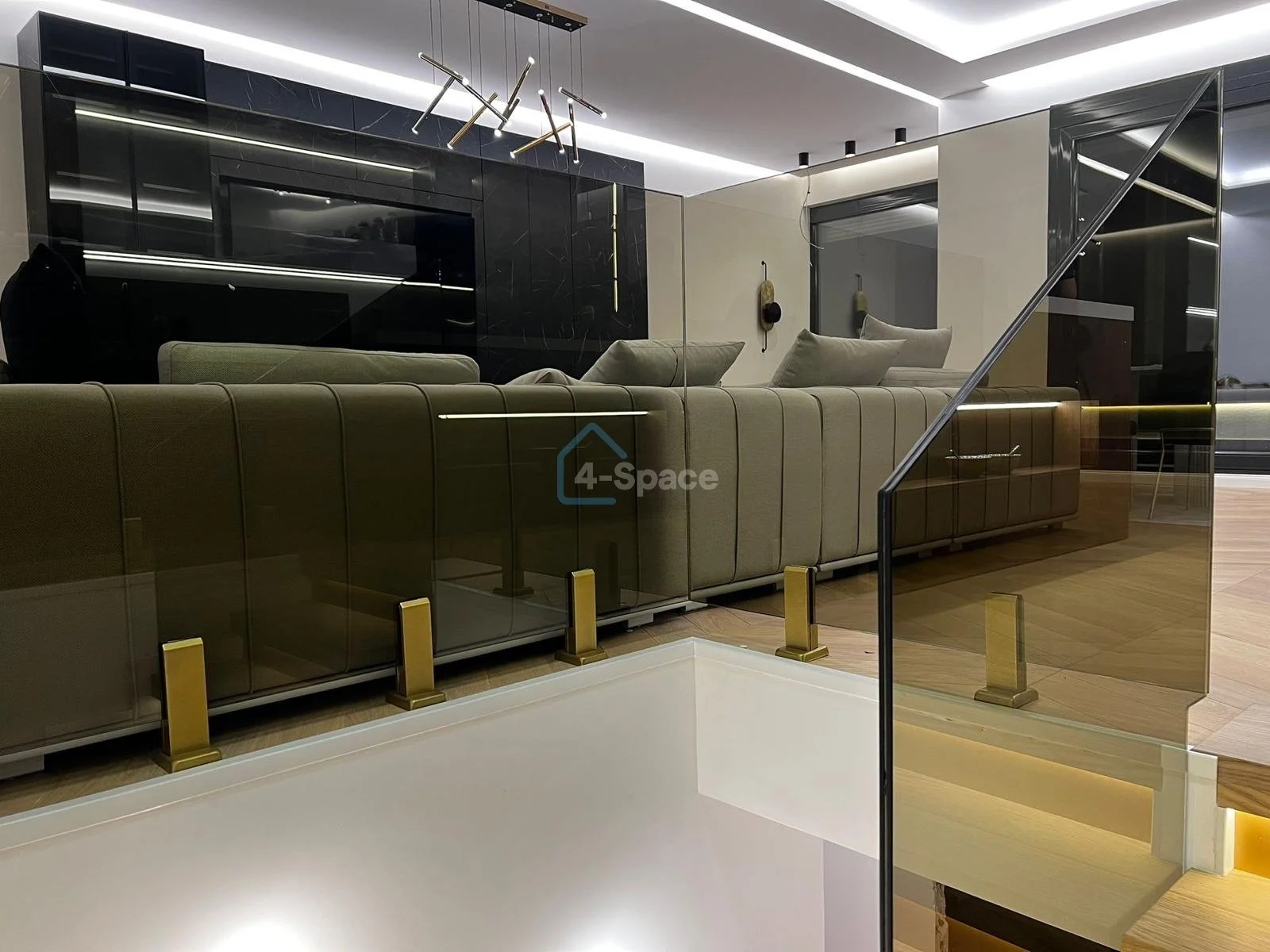 Interior of a modern living room featuring a beige sofa, glass railing, wall-mounted black entertainment center, and contemporary lighting fixtures.