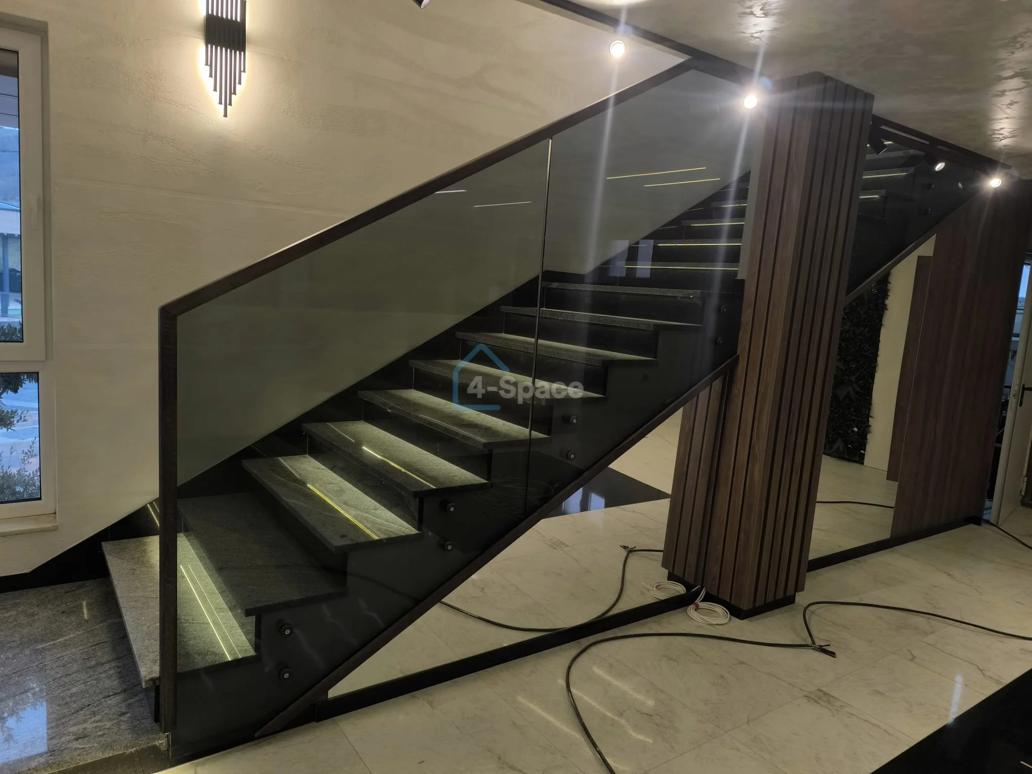 Modern indoor staircase with black steps, glass railing, and wooden panel accents, illuminated by ceiling lights.