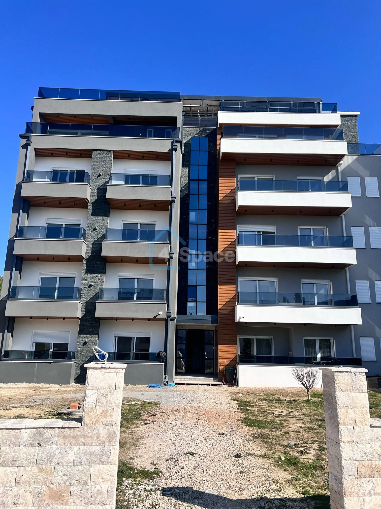 Modern multi-story apartment building with balconies and glass railings, surrounded by a partially constructed yard under a clear blue sky.