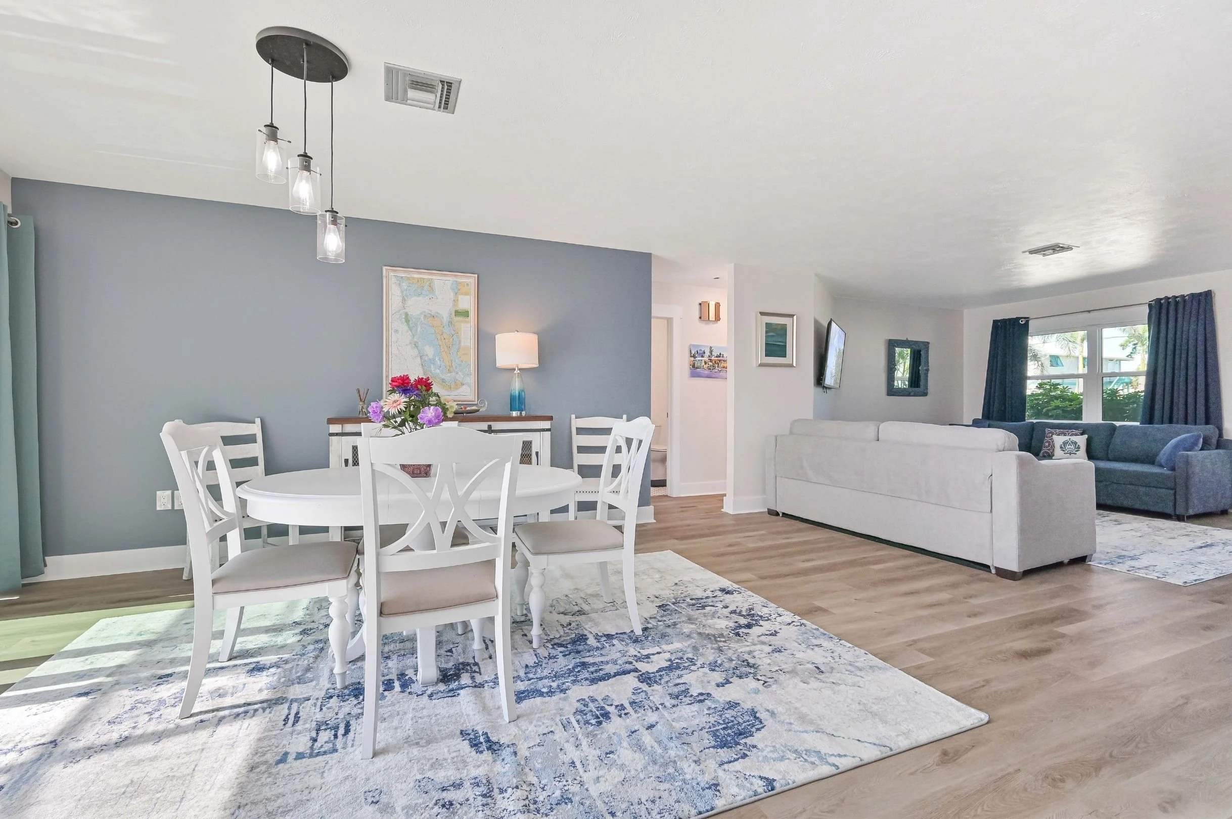 Open-concept living and dining area with light-colored hardwood floors, a round white dining table with six matching chairs, a floral centerpiece, a blue accent wall with a framed map, and a living room with white and blue sofas and windows with dark