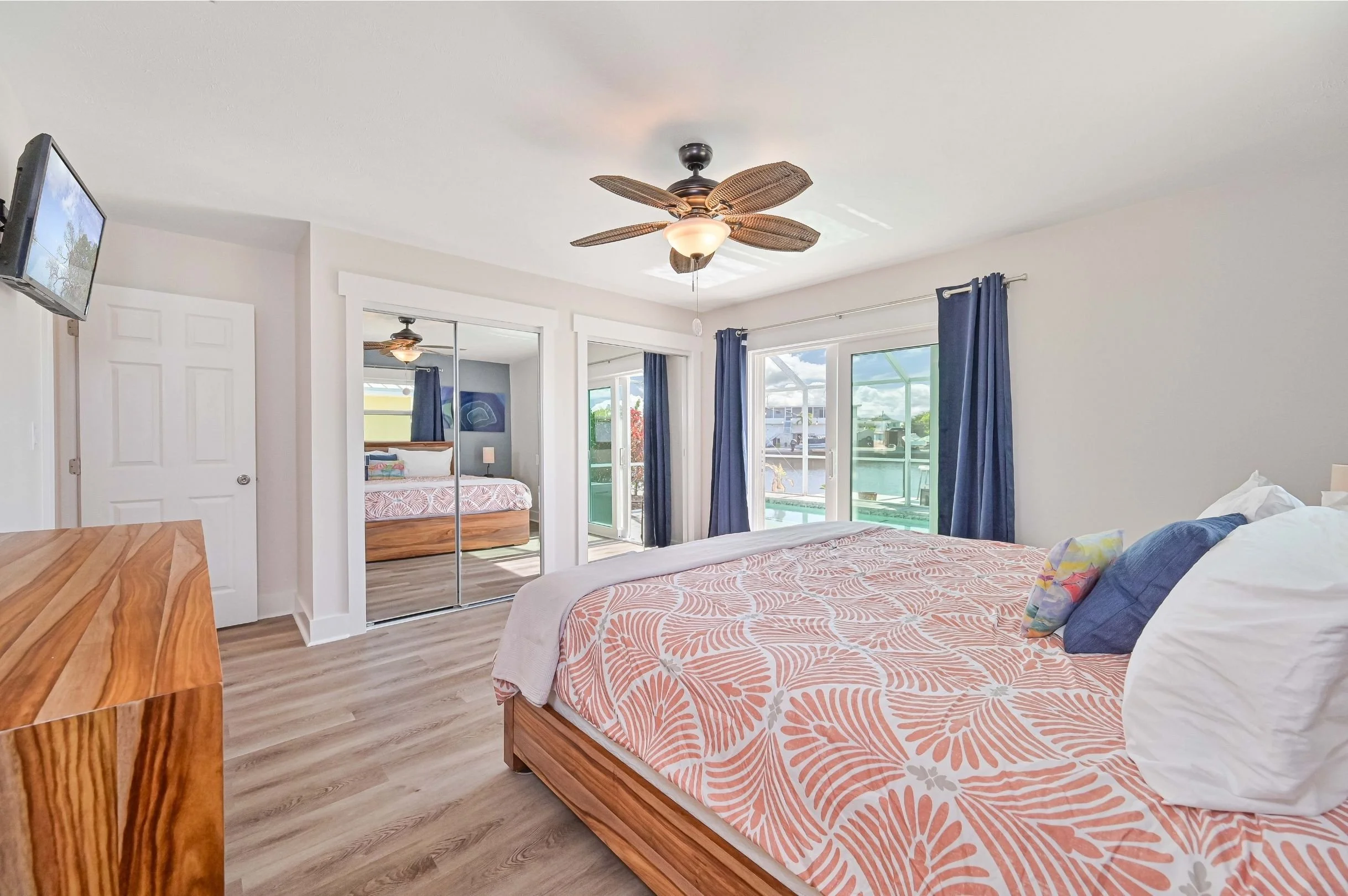 Bedroom with a wooden bed, patterned bedspread, white pillows, and dark blue pillows, a wooden dresser, mirrored closet doors, a ceiling fan with palm leaf design, and large sliding glass doors with blue curtains leading to a screened outdoor area wi