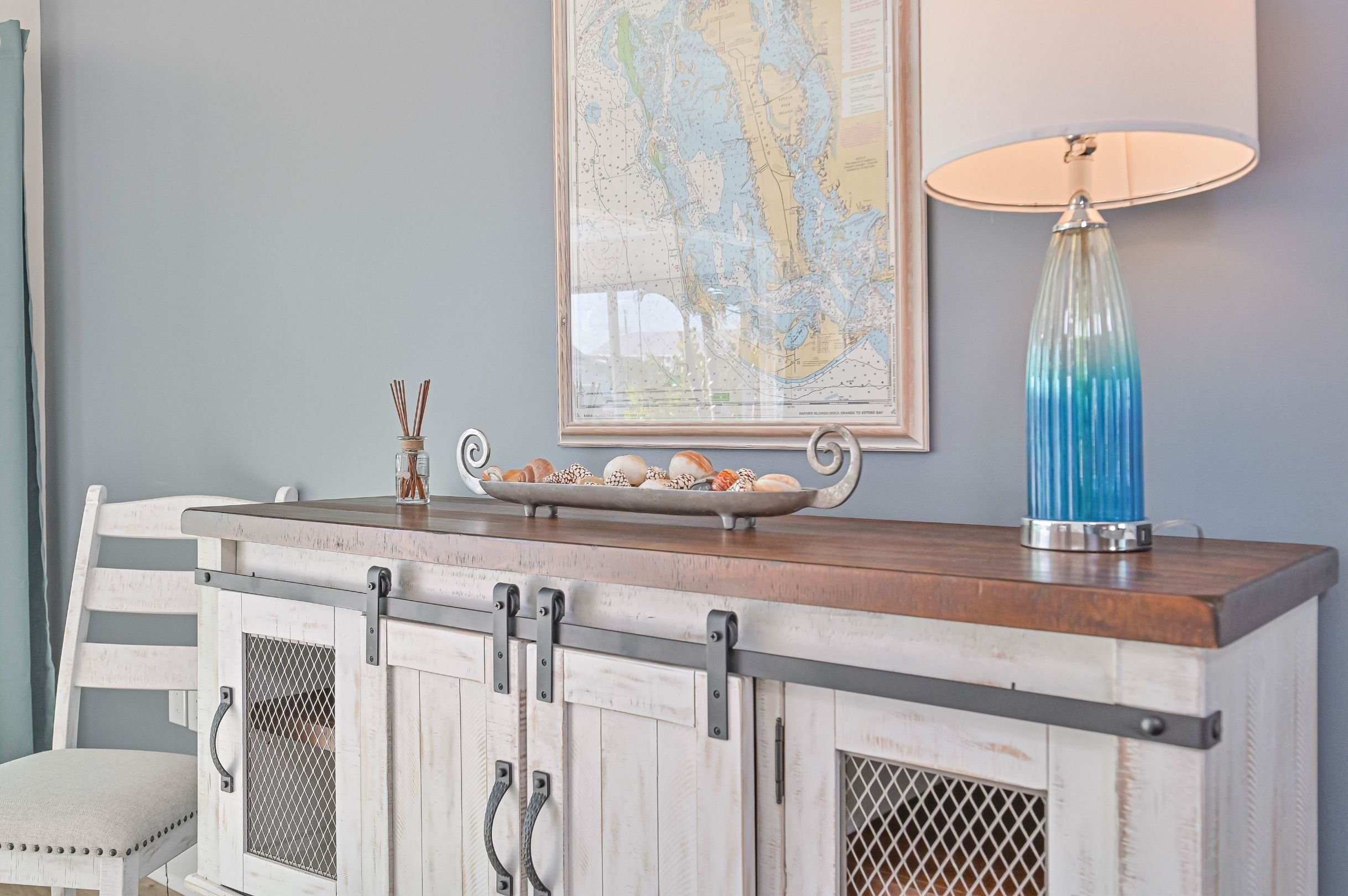 A wooden sideboard with a white painted finish, sliding barn door hardware, and mesh panels, topped with a decorative tray of seashells, a bottle of reed diffuser, and a large table lamp with a blue gradient base and white shade, against a light blue