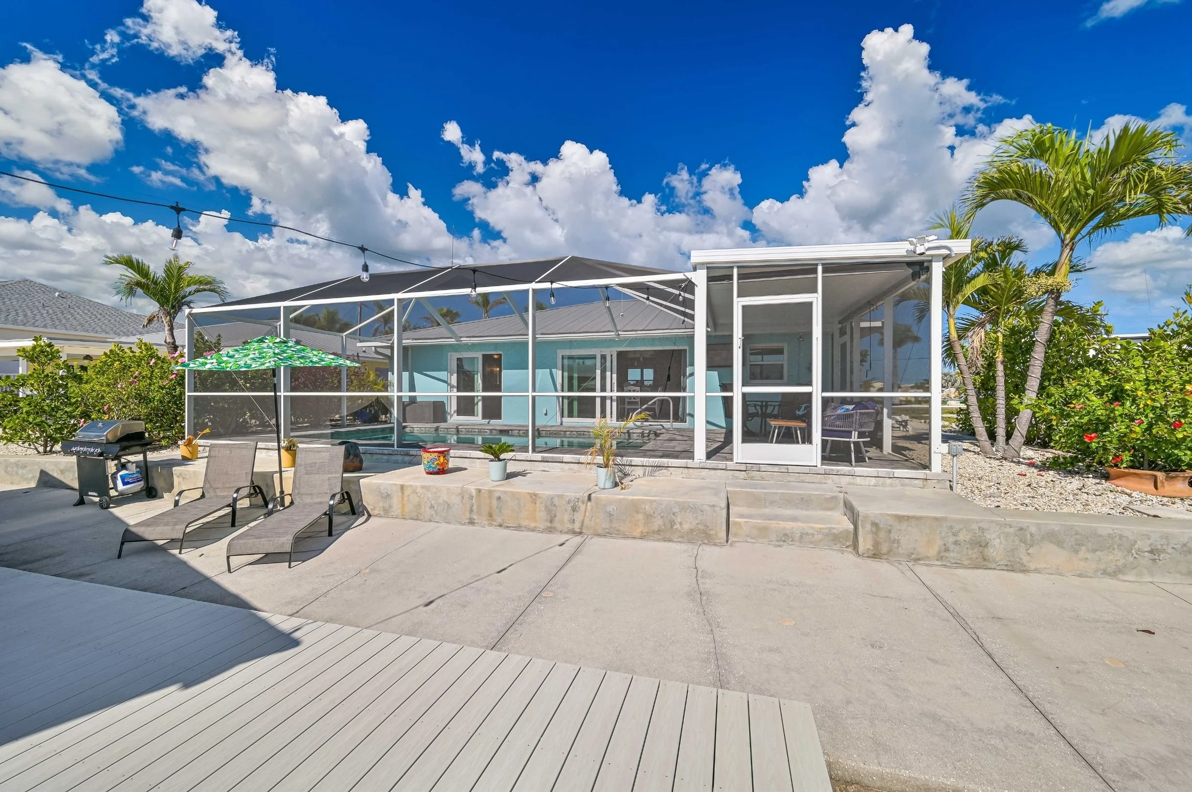 Backyard patio with a screened porch, two lounge chairs, a grill, and tropical plants under a blue sky with scattered clouds.