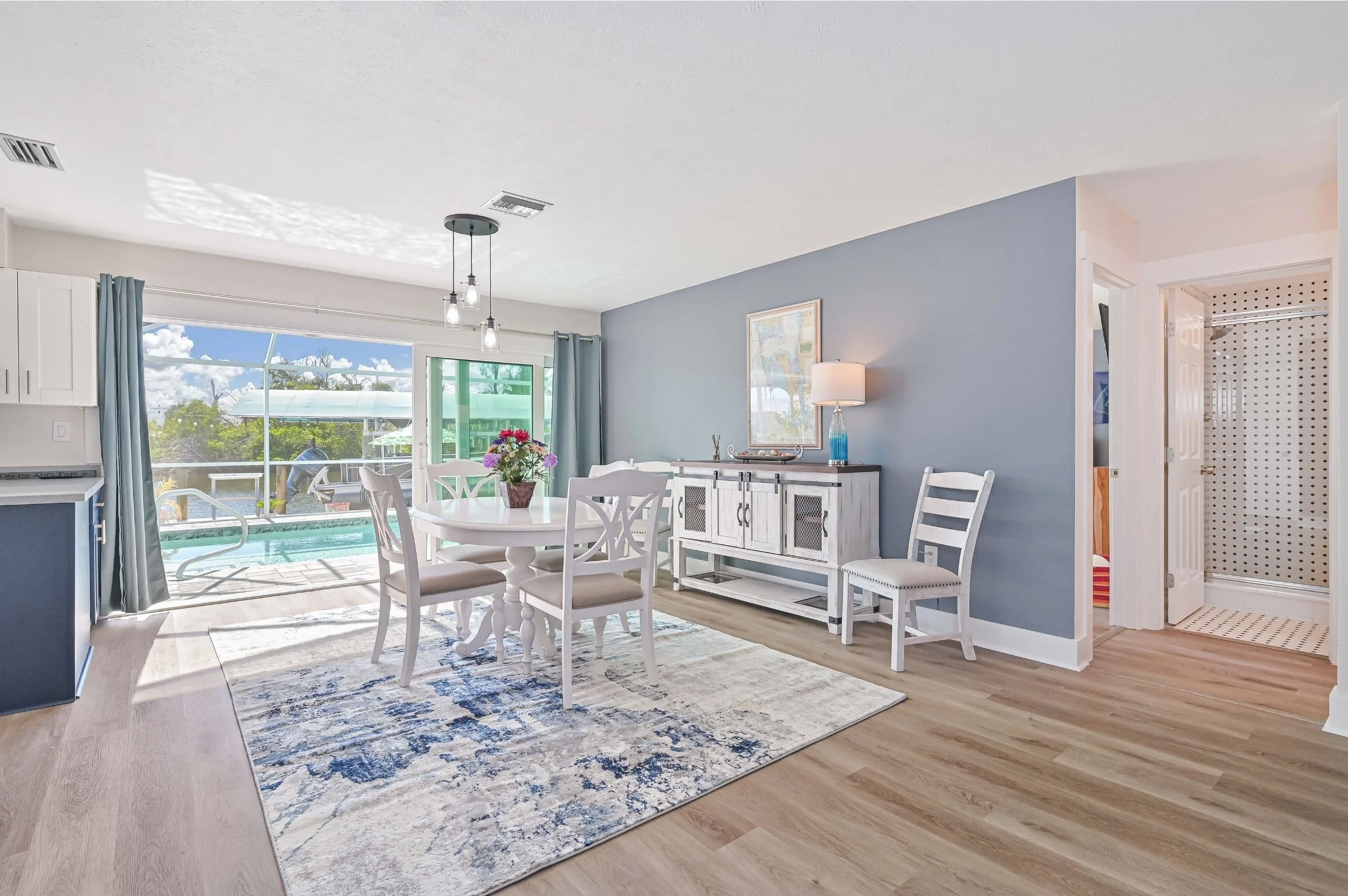 Bright dining area with a round white table and six chairs, a white sideboard with a lamp on it, a sliding glass door leading to a pool, and a blue accent wall.