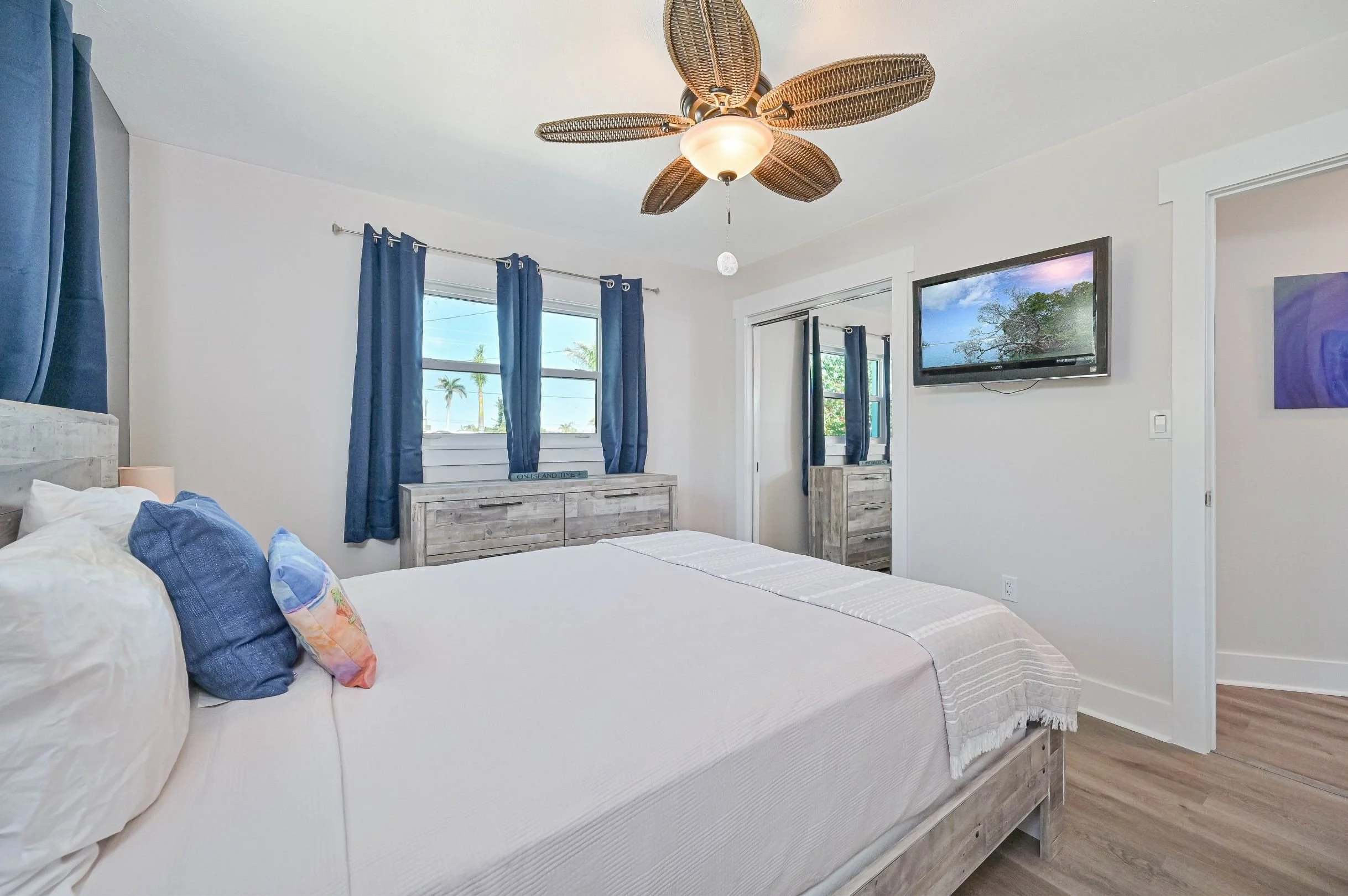 Bright bedroom with white walls, a wooden bed frame, blue curtains, a ceiling fan with leaf-shaped blades, a wall-mounted TV, and wooden dressers, with windows showing palm trees outside.