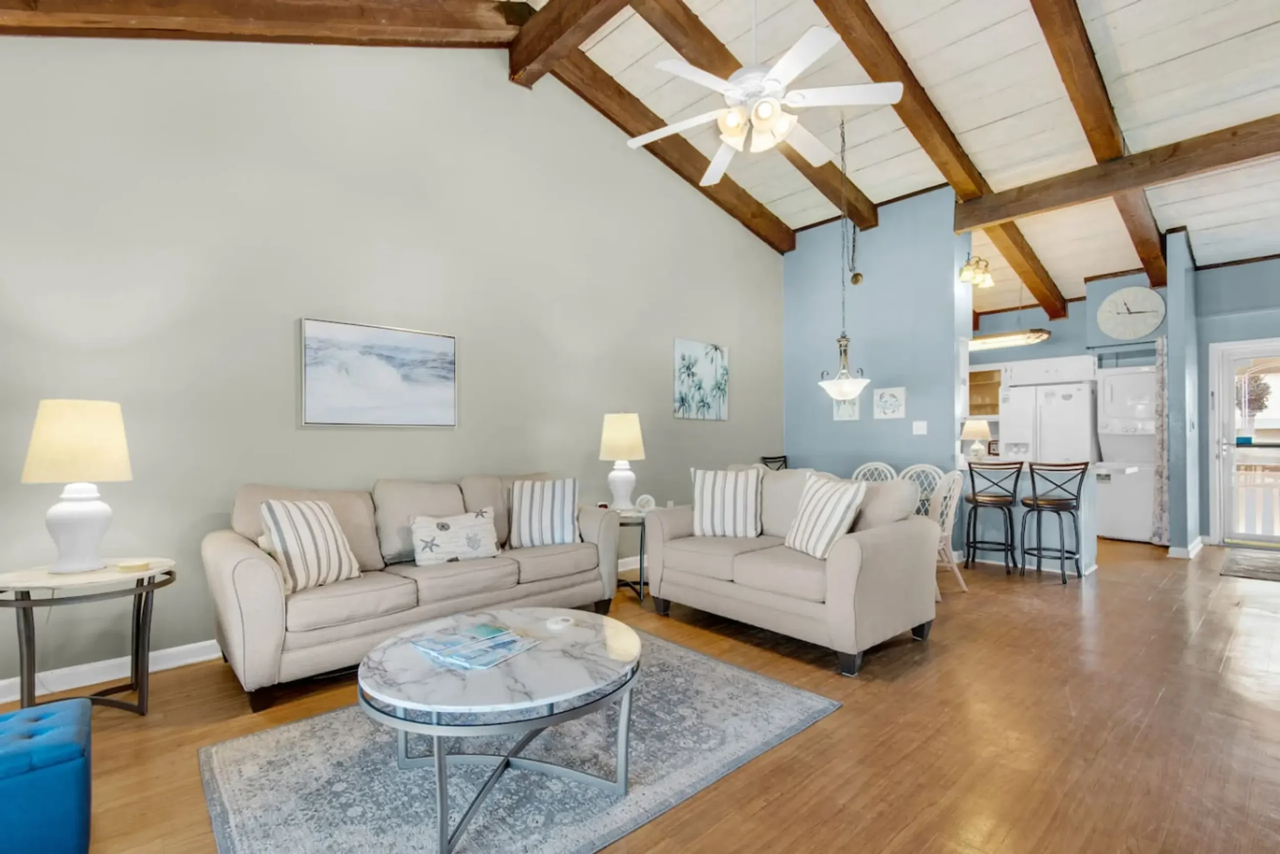 Living room with light-colored sofas, wooden ceiling with exposed beams, a marble coffee table, and a blue accent wall with artwork, overlooking a kitchen area with bar stools and a refrigerator.