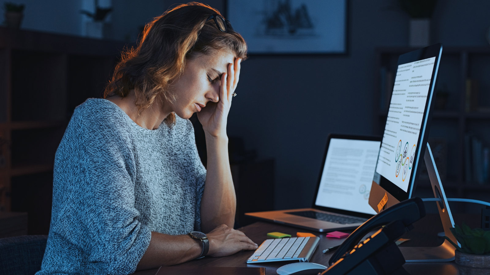 Woman with her eyes closed, head in hand in dark home office desktop laptop and landline.