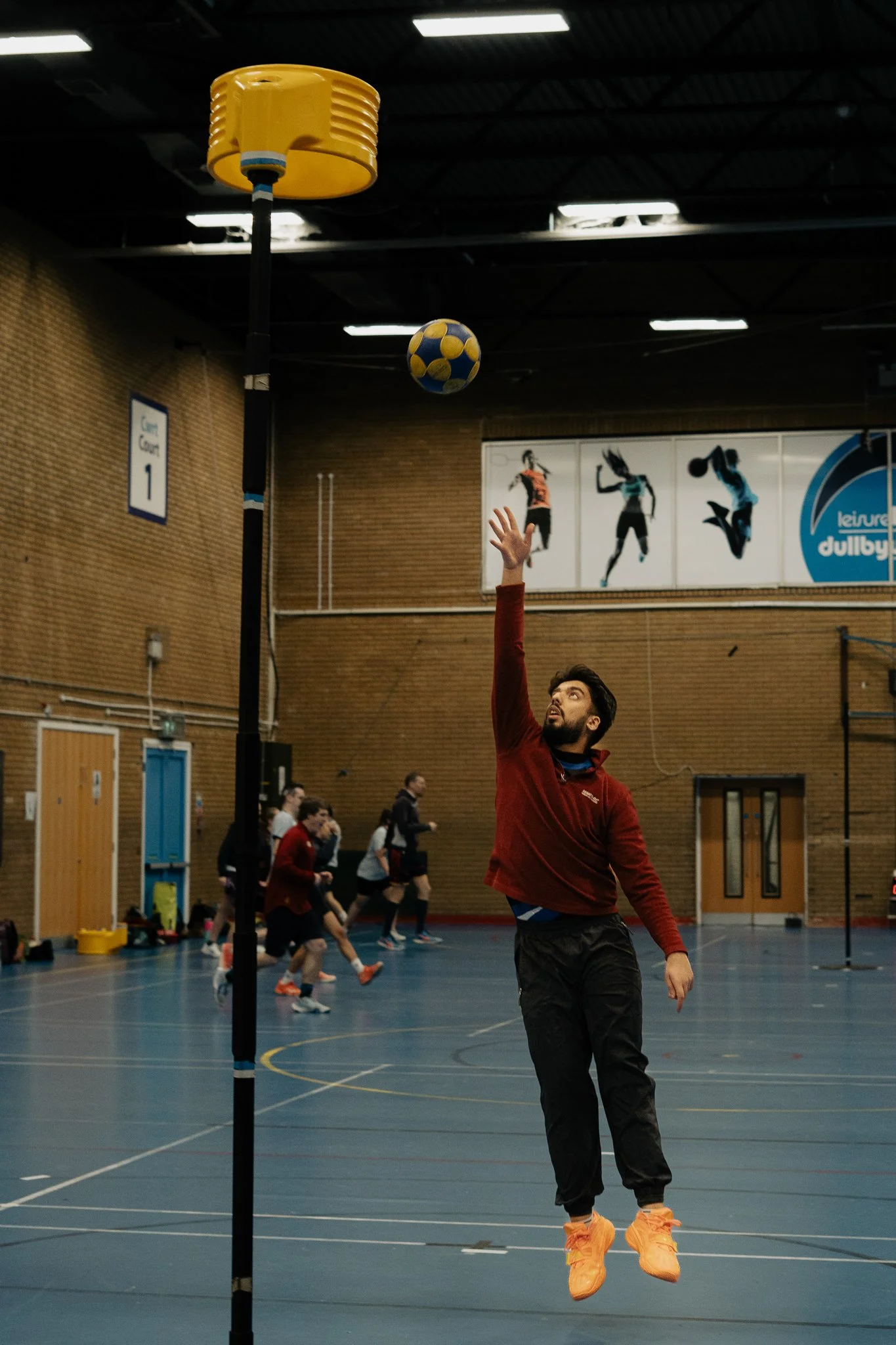 A man in a red jacket and black pants jumping to hit a volleyball in an indoor sports gymnasium.