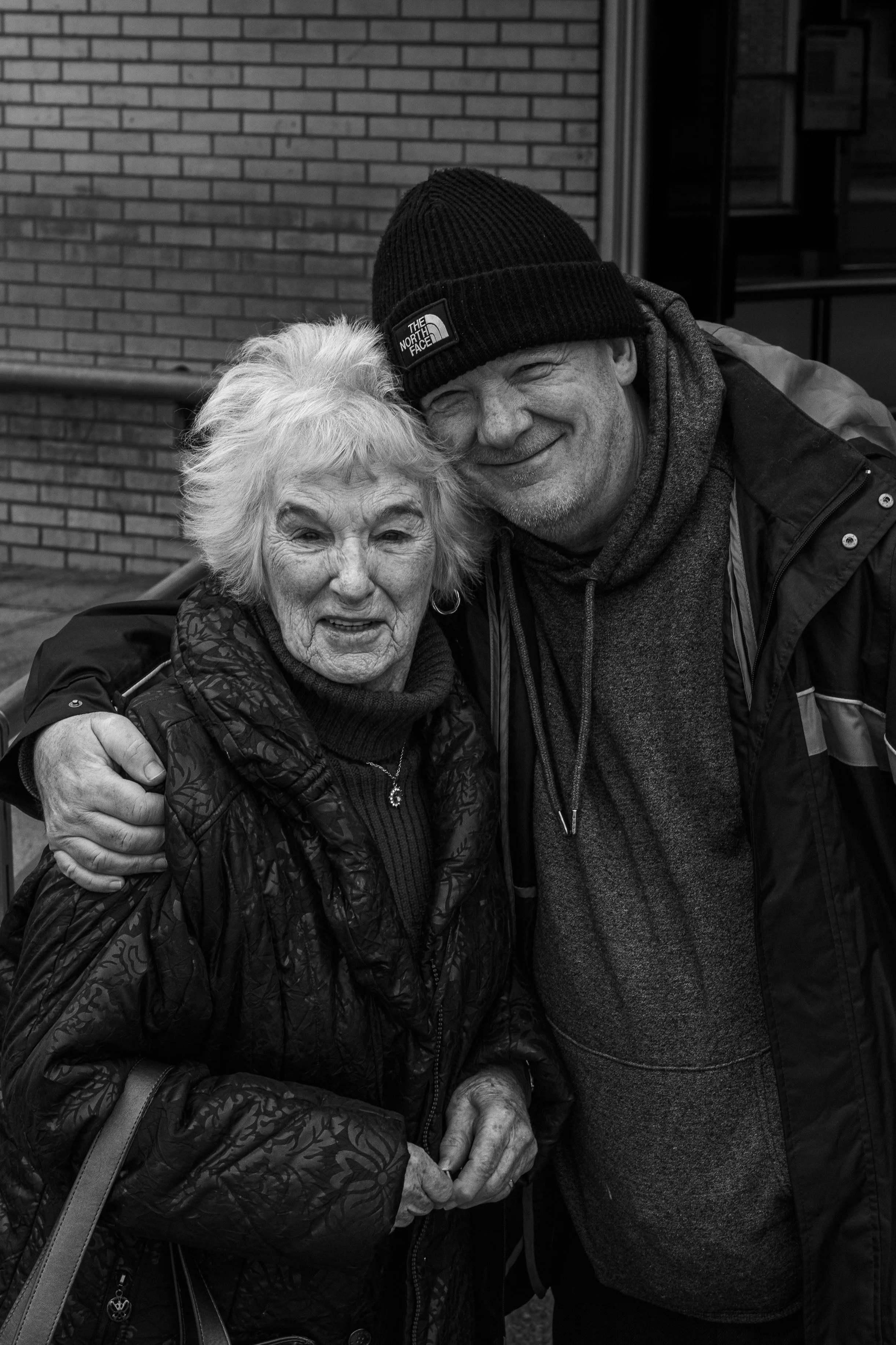 A black and white photo of a smiling elderly woman and a smiling middle-aged man, hugging each other outdoors, in front of a brick wall. The man is wearing a black beanie, hoodie, and jacket. The woman has light-colored hair and is wearing a patterne