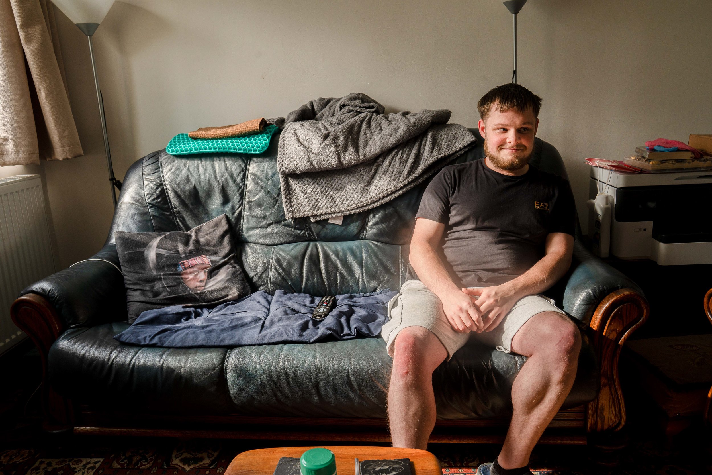 A young man with a beard sitting on a black leather sofa, smiling, with clutter on the backrest, including blankets and a pillow with a face on it, in a living room with standing lamps and a side table.