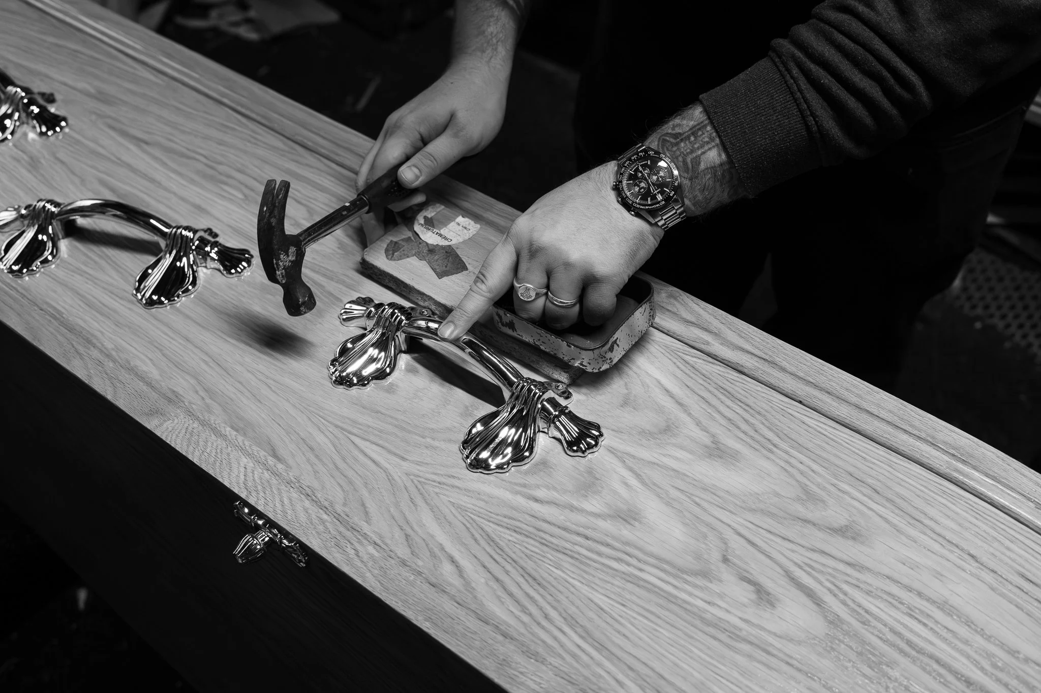 A person working on a jewelry piece, using a hammer and a small chisel on a wooden workbench, with shiny metallic jewelry pieces laid out.
