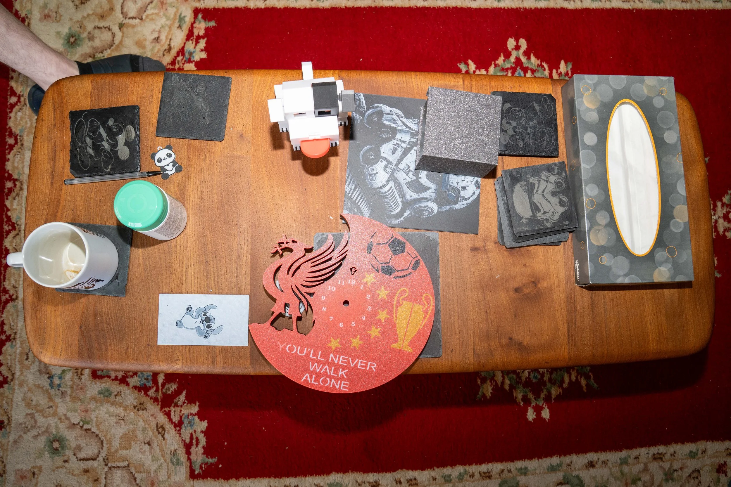 Top-down view of a wooden table with various items including a cup, a tissue box, a small robotic figure, a keychain, black pieces, a red Liverpool football club mascot cutout with the phrase "You'll never walk alone," framed artwork, a black and whi
