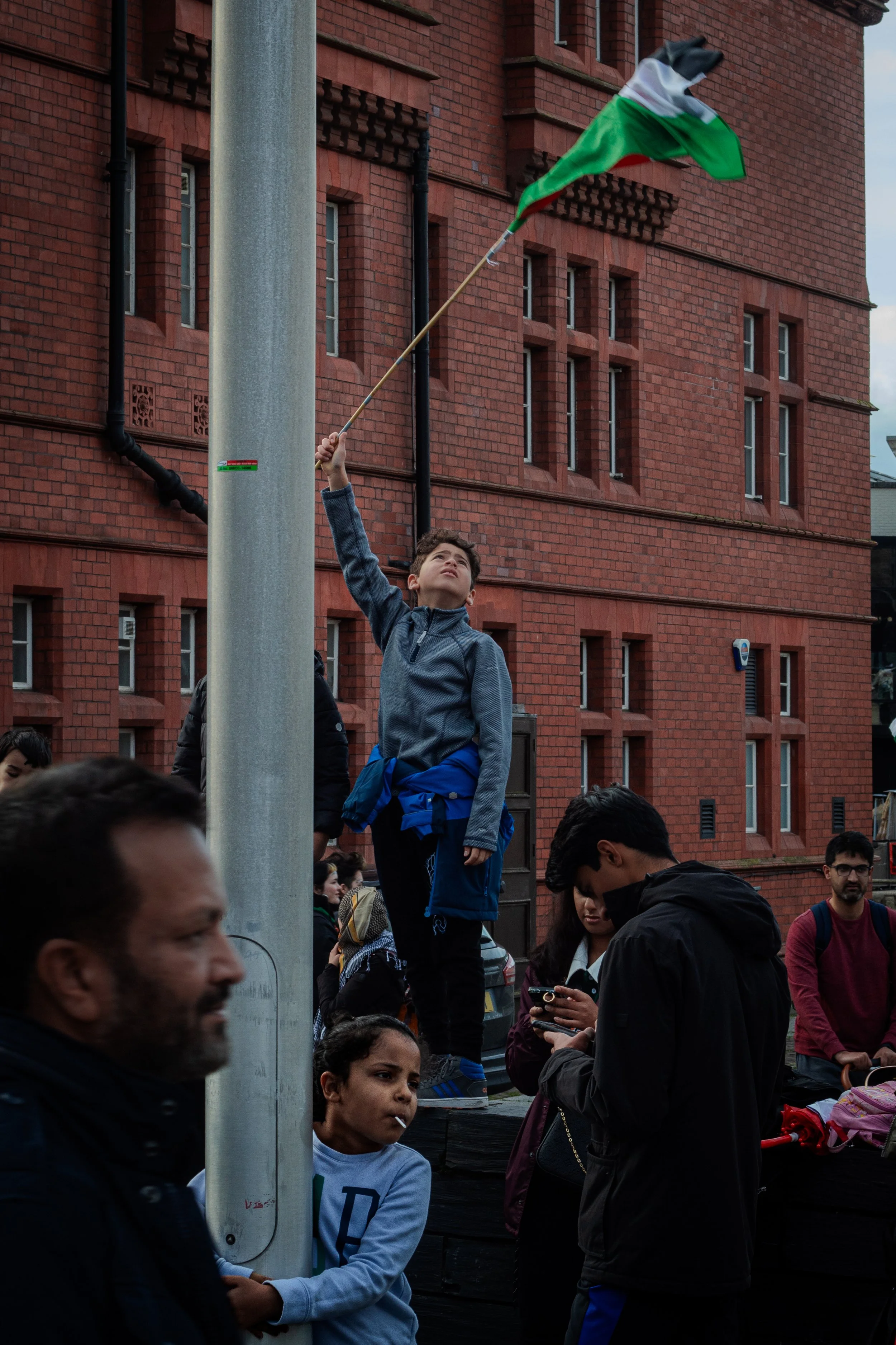 A boy standing on a platform raising a flagpole with a green, white, and black flag against a red brick building, with several people around, some using phones.