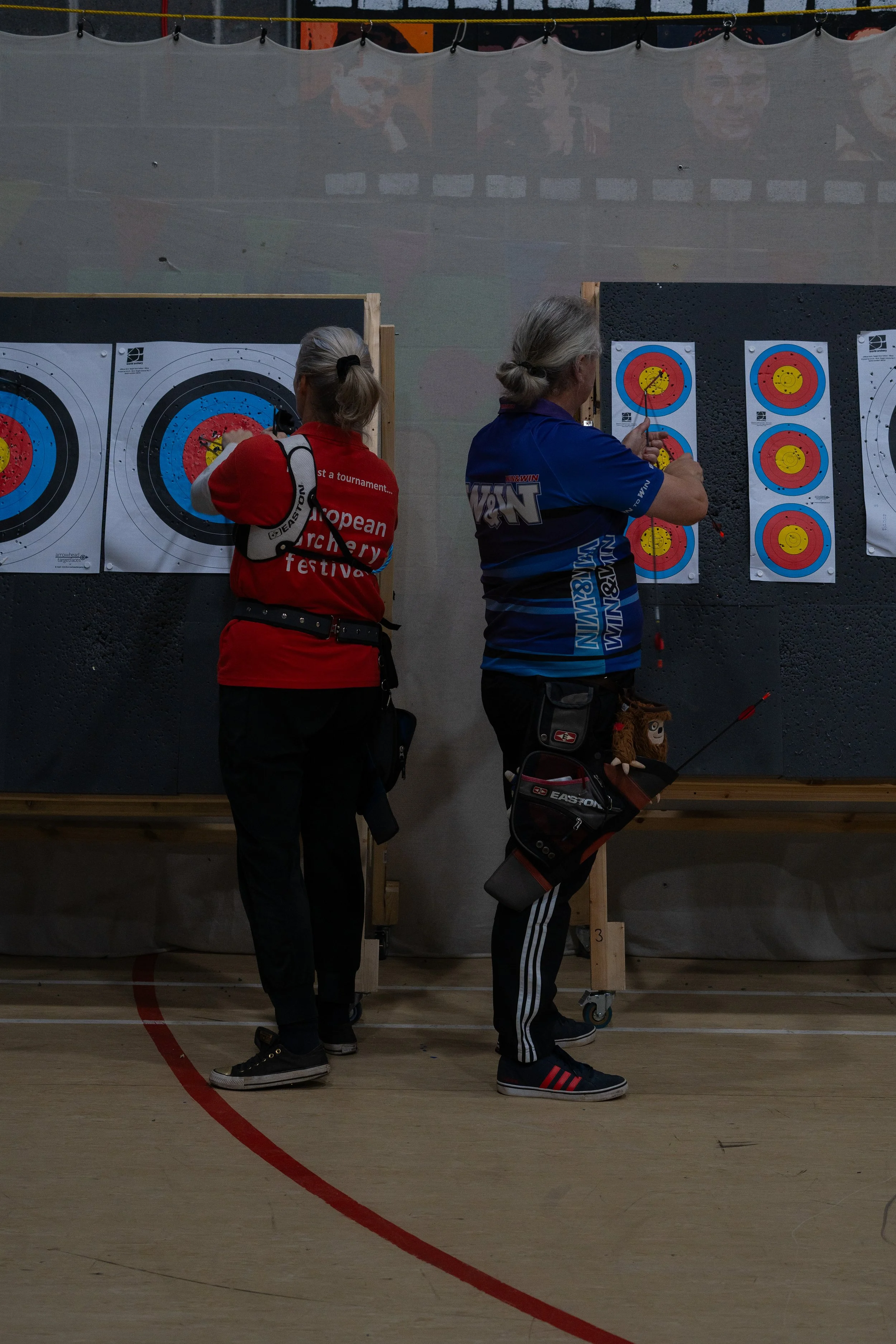 Two women archers aiming at targets with arrows at an indoor archery range.