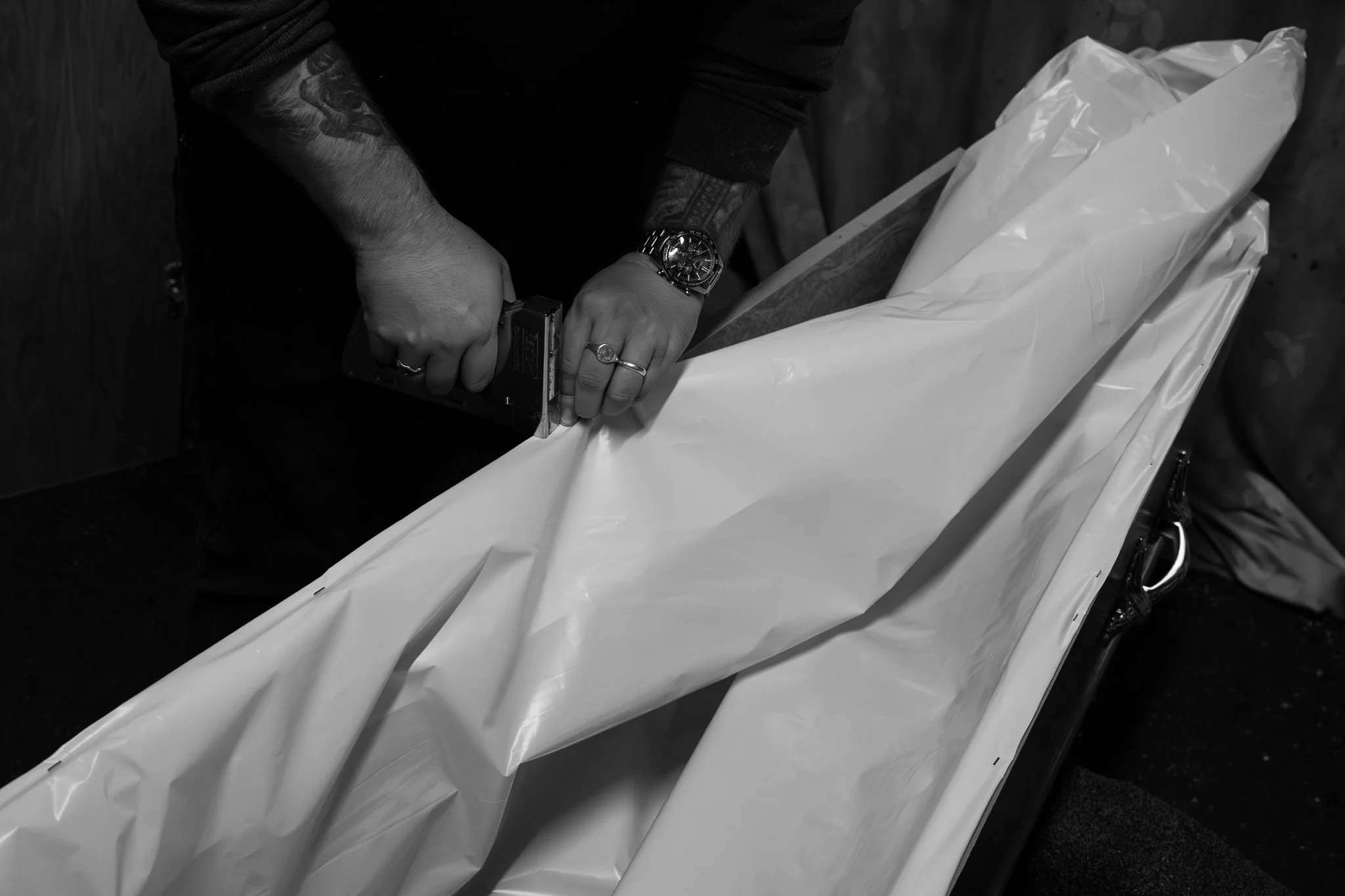 A person is opening a body bag with a pair of hands visible, wearing a wristwatch and rings. The person's arms are tattooed. The body bag is on a table or stretcher, and the background is dark.