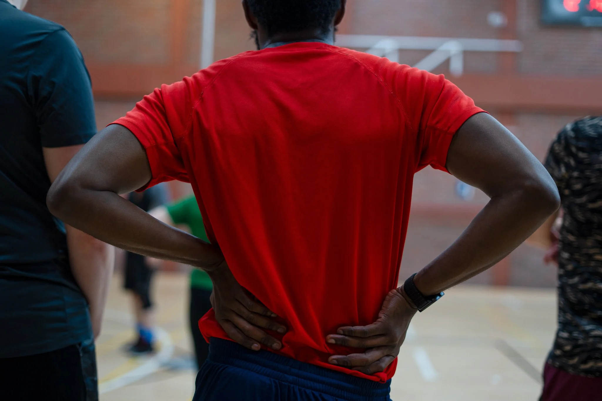 Person in red athletic shirt with hands on hips in a gymnasium or sports hall.