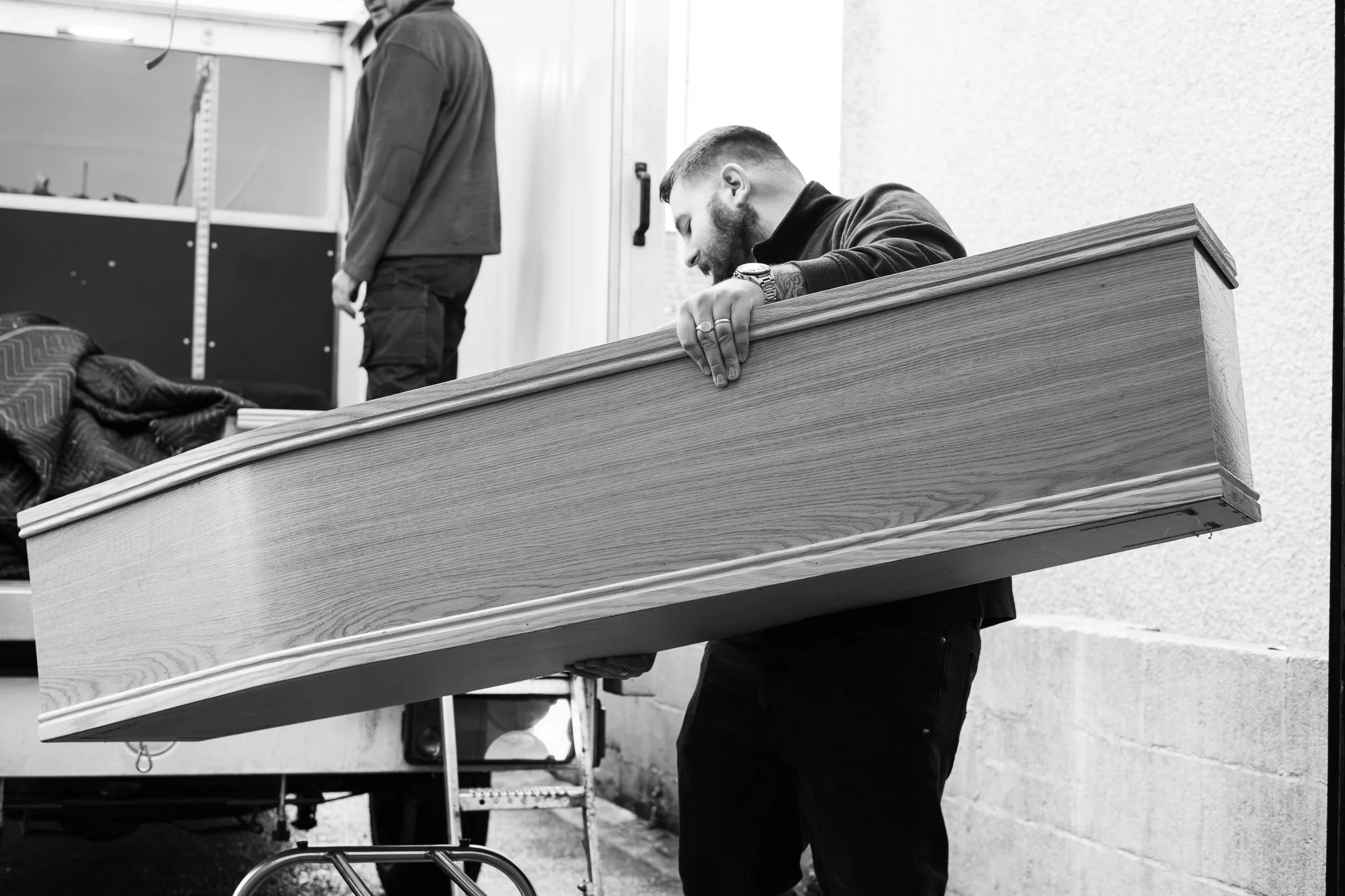 A man in dark clothing carrying a large wooden casket, with another person in the background, inside a workshop or storage area.