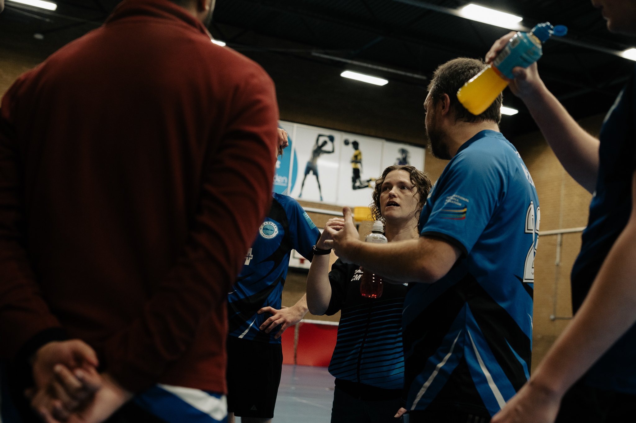 A group of athletes and a coach having an intense discussion during a break in an indoor sports facility. The woman in the center appears to be listening attentively, holding a water bottle. The surroundings include sports posters on the wall.