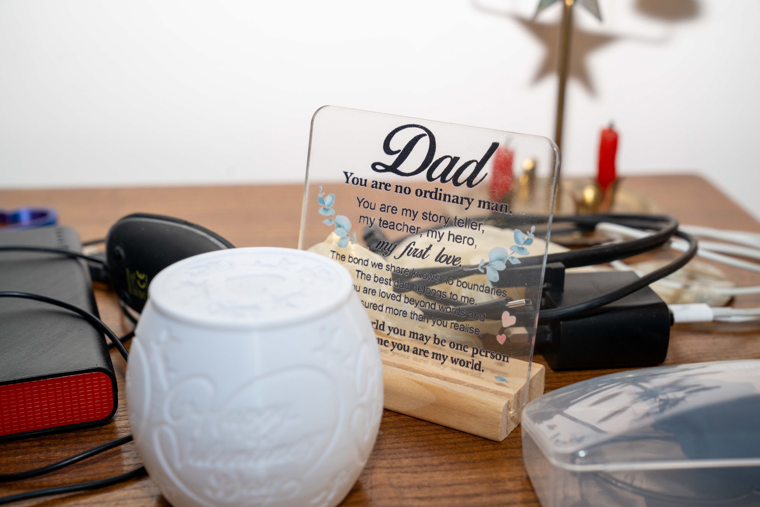 A clear acrylic plaque with a loving message for Dad, placed on a wooden desk among electronic devices, a white decorative pot, and a plastic container.